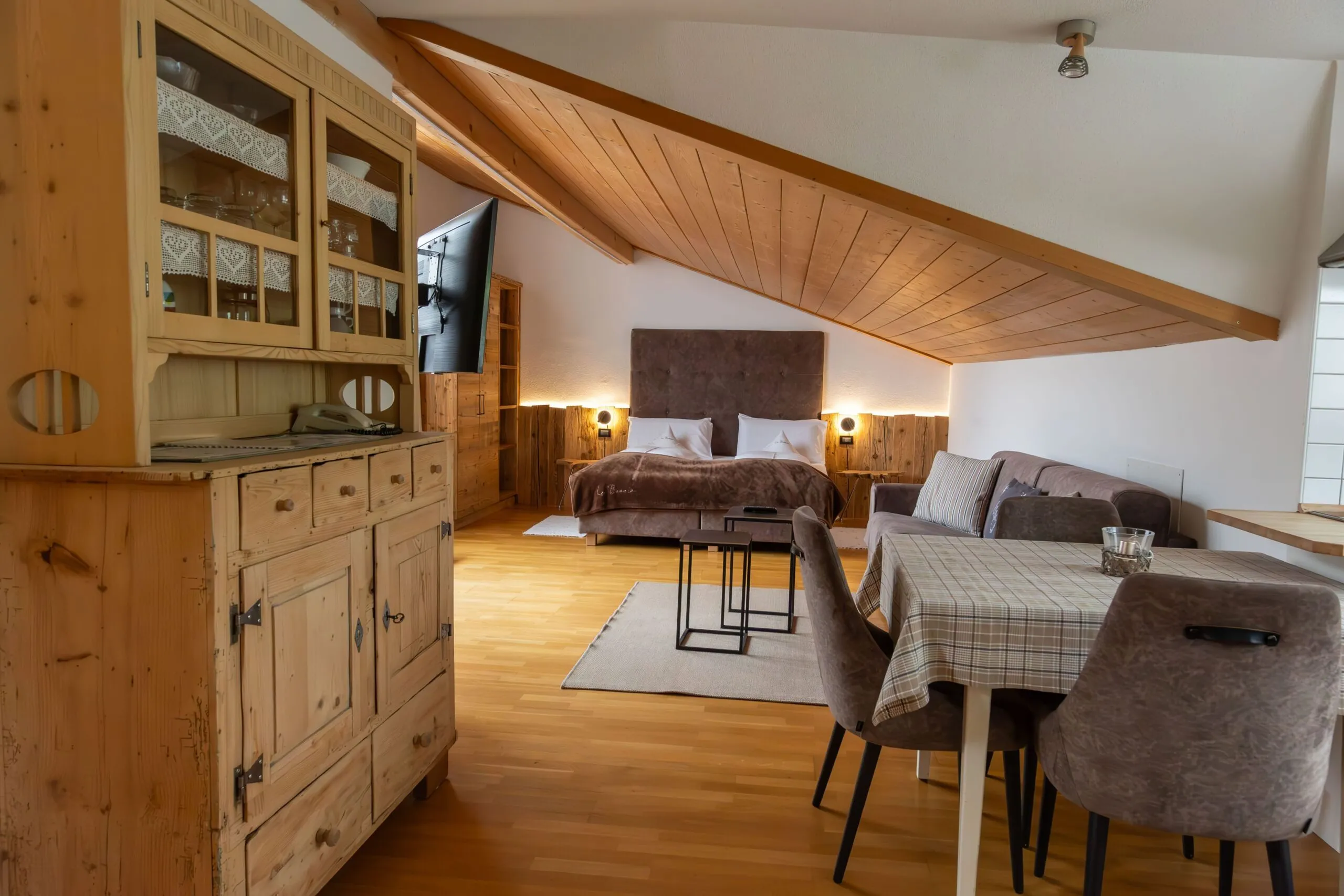 Cozy alpine living area with a wooden cabinet, slanted wooden ceiling, bed, seating, and dining table.