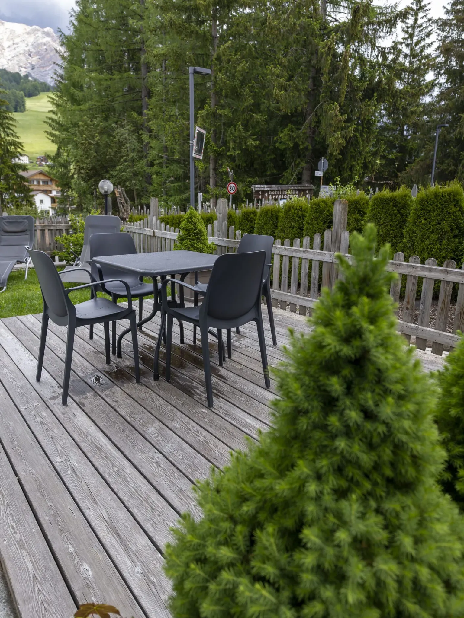 Outdoor wooden deck with gray plastic table and chairs, surrounding hedge and trees in a mountain setting.