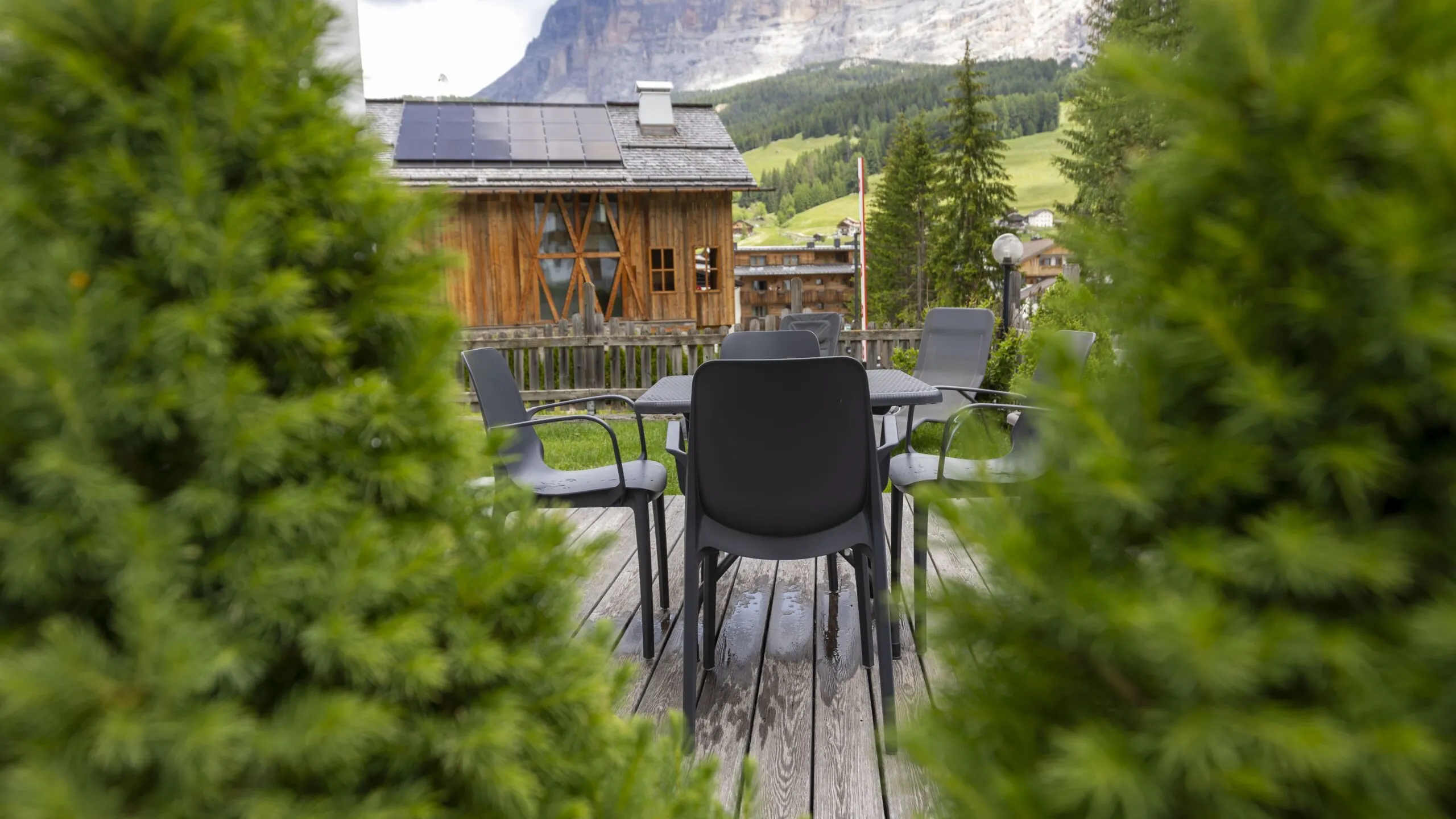 Outdoor wooden terrace with gray chairs and table, rustic cabin, green hills, and mountains in the background.