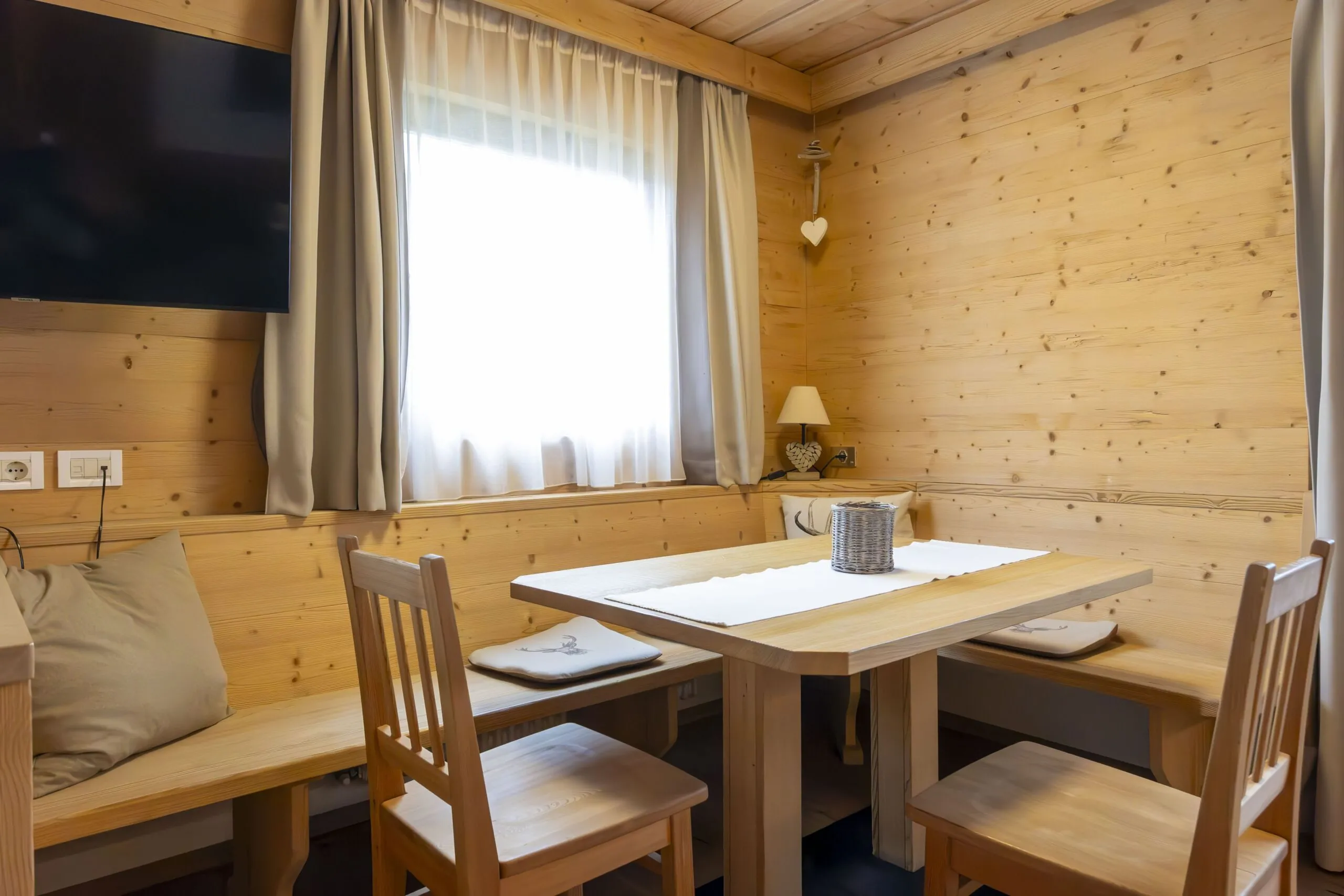 Cozy wooden dining nook with a rectangular table, bench seating, chairs, and a window with sheer white curtains.