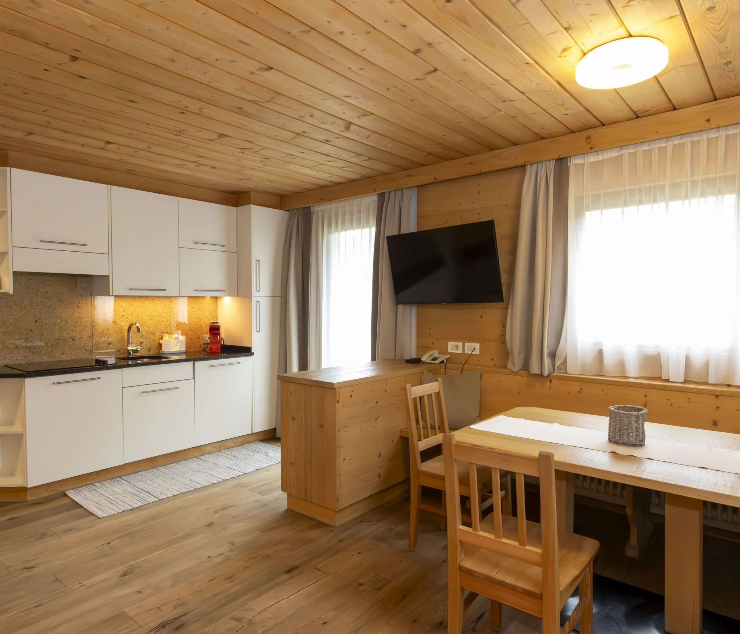 Cozy wooden kitchen-dining area with white cabinets, granite backsplash, and a wall-mounted TV by a window with light curtains.