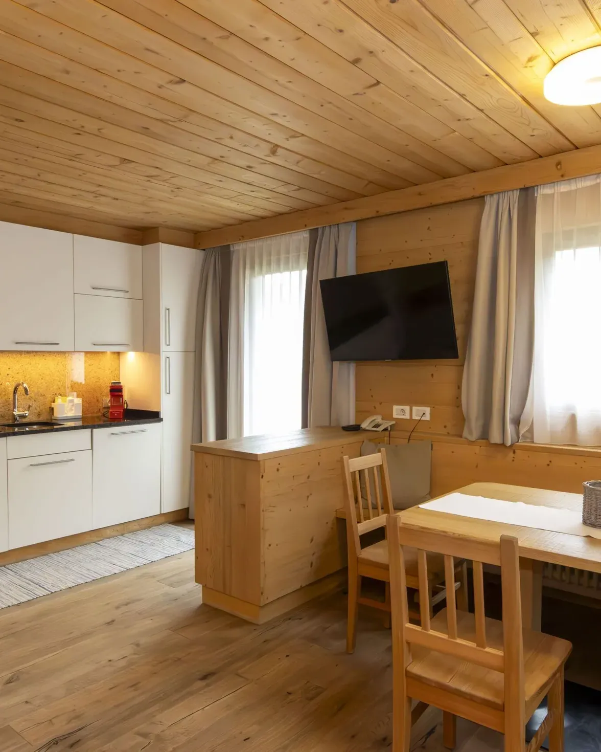 Cozy wooden kitchen-dining area with white cabinets, granite backsplash, and a wall-mounted TV by a window with light curtains.