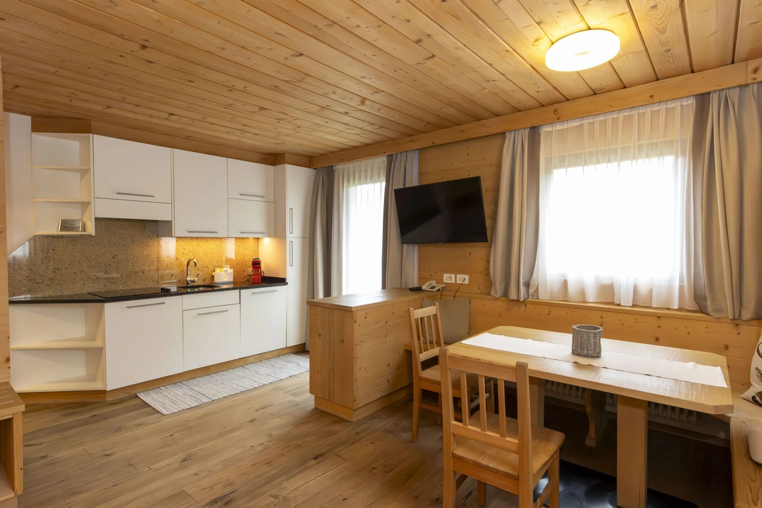 Cozy wooden kitchen-dining area with white cabinets, granite backsplash, and a wall-mounted TV by a window with light curtains.