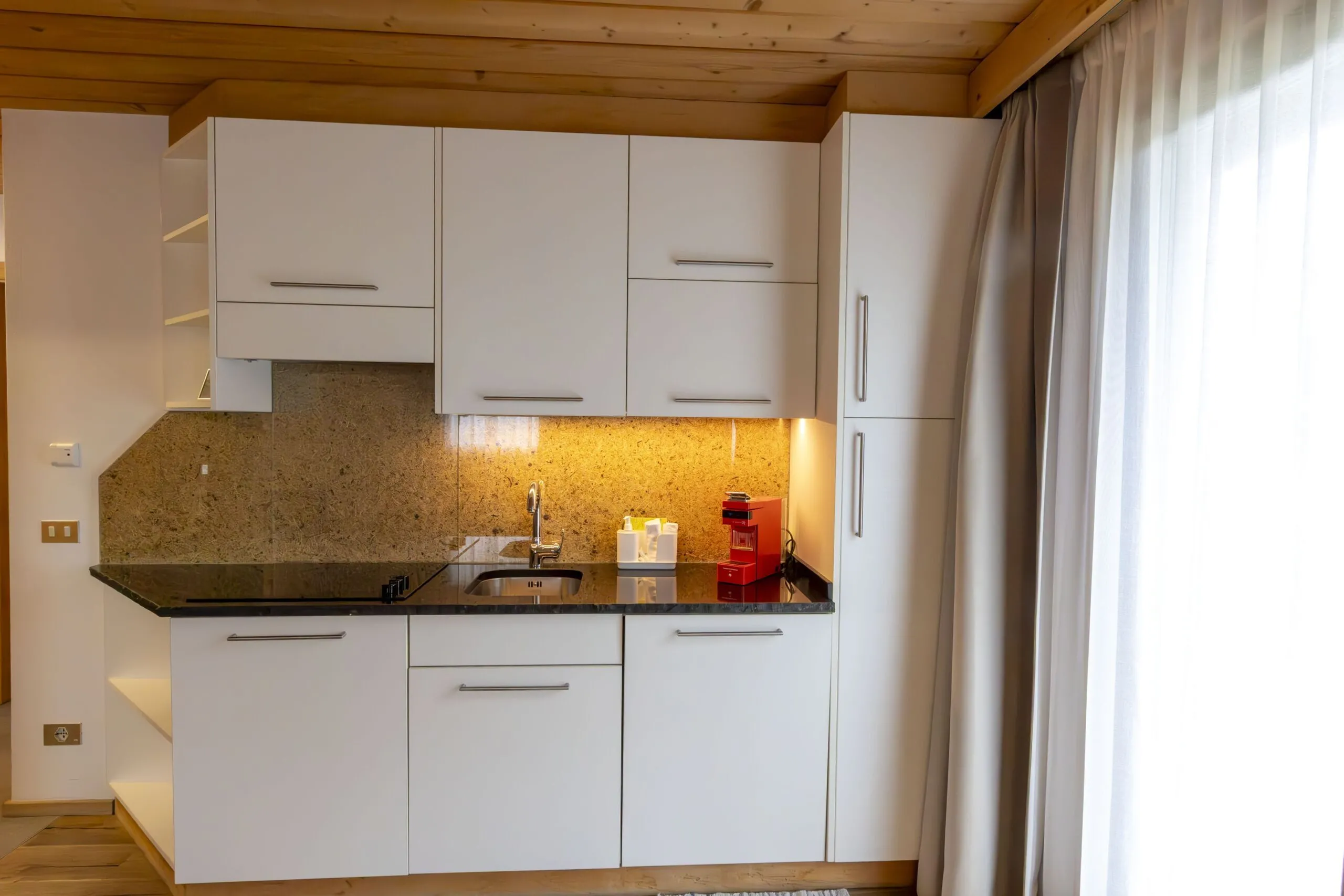 Modern white kitchen with gray granite countertop, built-in cabinets, sink, and small red coffee machine by window curtain.