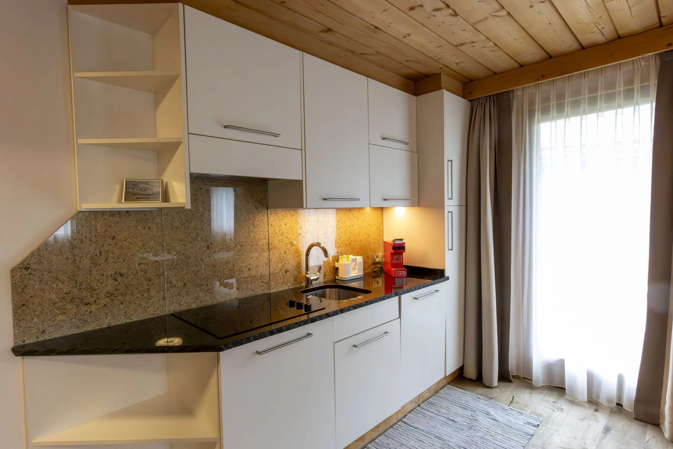 Modern white kitchen with granite countertop and backsplash, cabinets, sink, and a red coffee machine near a large window with sheer curtains.
