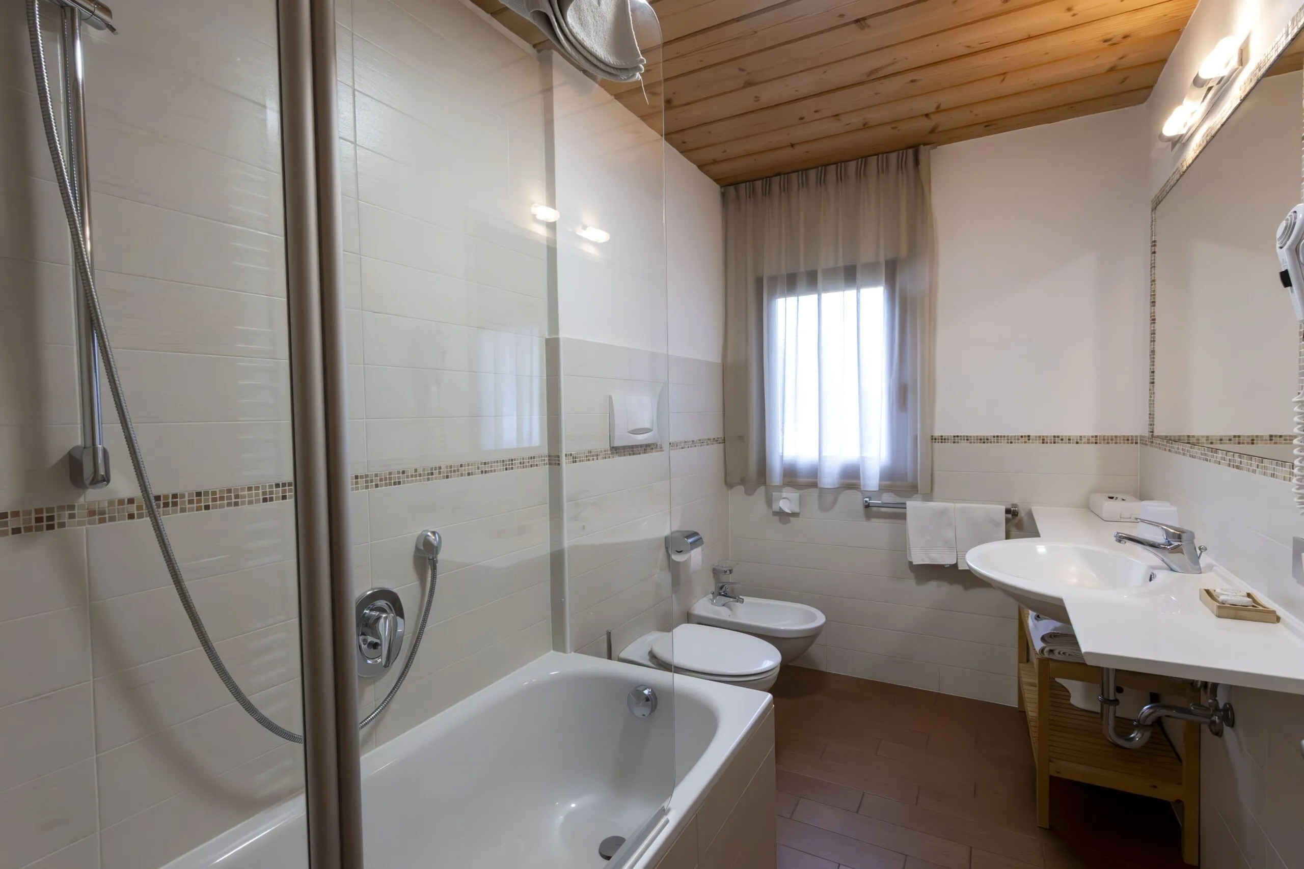 Bright bathroom with white tiled walls, wooden ceiling, glass shower, bathtub, and sink area.