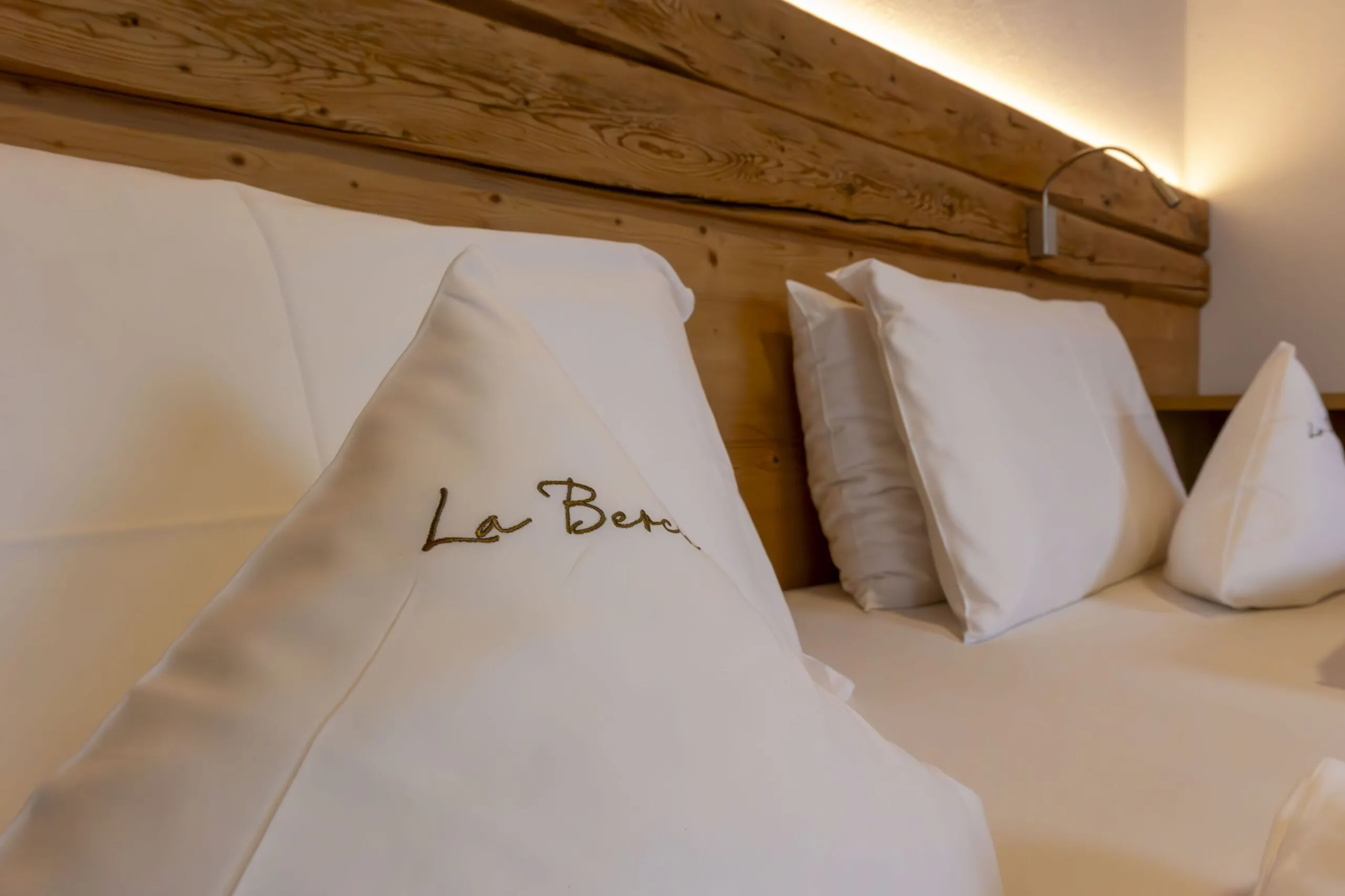 Luxurious hotel bed with white pillows, embroidered “La Ber” detail, wooden headboard, warm ambient lighting