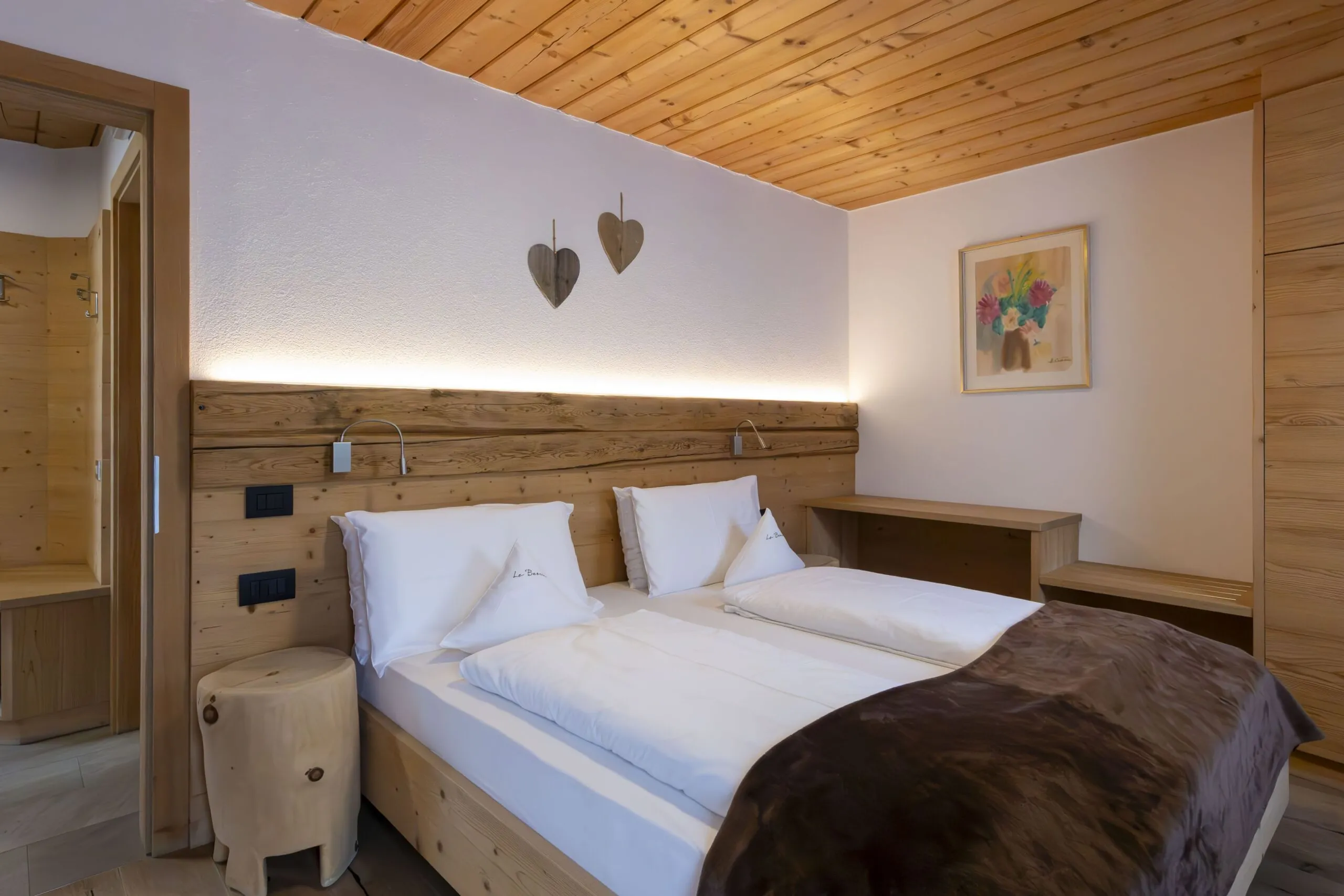 Cozy wooden hotel room with two beds, white bedding, brown blanket, warm wall lights, heart decor, and bedside desk.