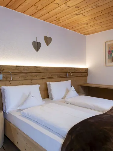 Cozy wooden hotel room with two beds, white bedding, brown blanket, warm wall lights, heart decor, and bedside desk.