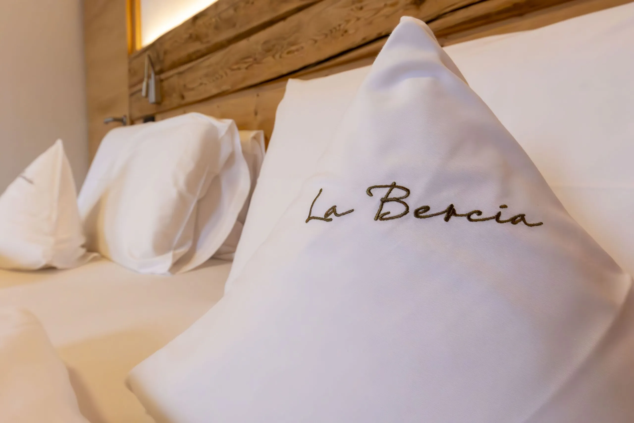 Luxurious hotel bed with white pillowcase embroidered "La Beria" in brown thread, wood headboard in background.
