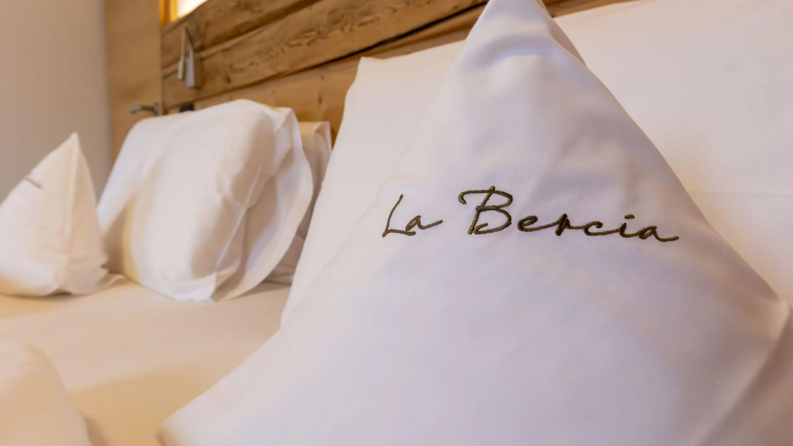 Luxurious hotel bed with white pillowcase embroidered "La Beria" in brown thread, wood headboard in background.