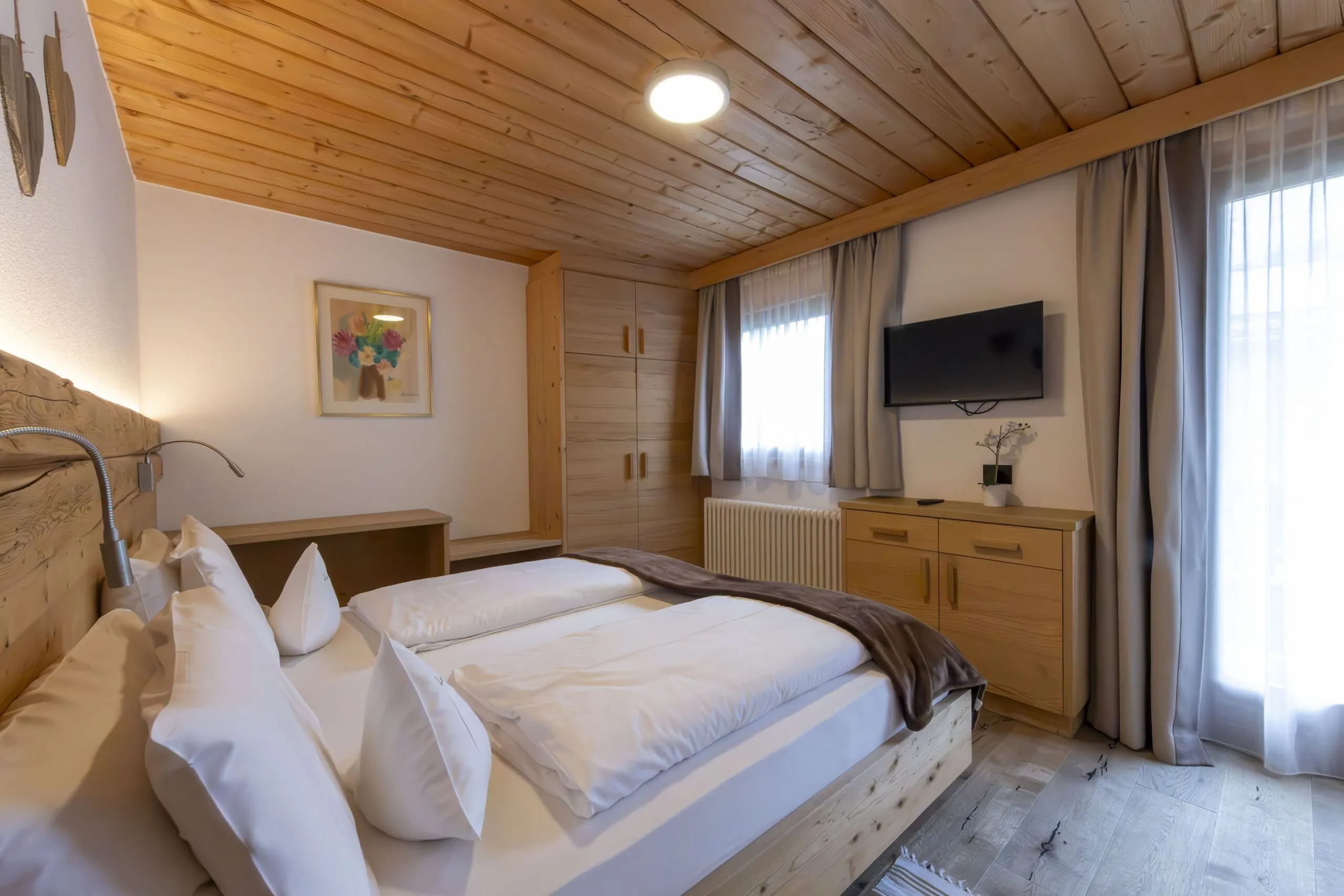 Cozy wooden bedroom with a large bed, bedside lamp, wardrobe, dresser, and wall-mounted TV under a paneled ceiling.