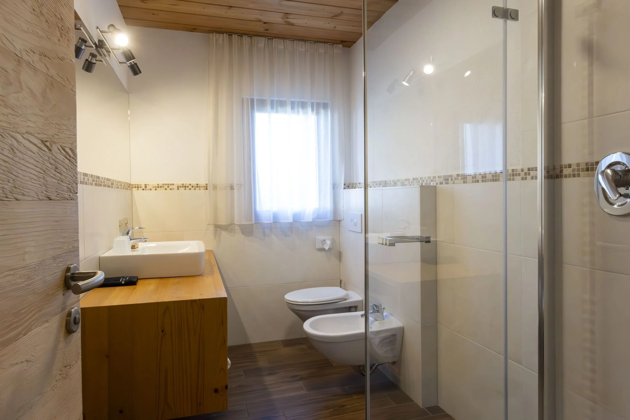 Bright modern bathroom with wooden vanity, rectangular sink, window with sheer curtains, and wall mosaic tile accent; glass shower enclosure, toilet and bidet.