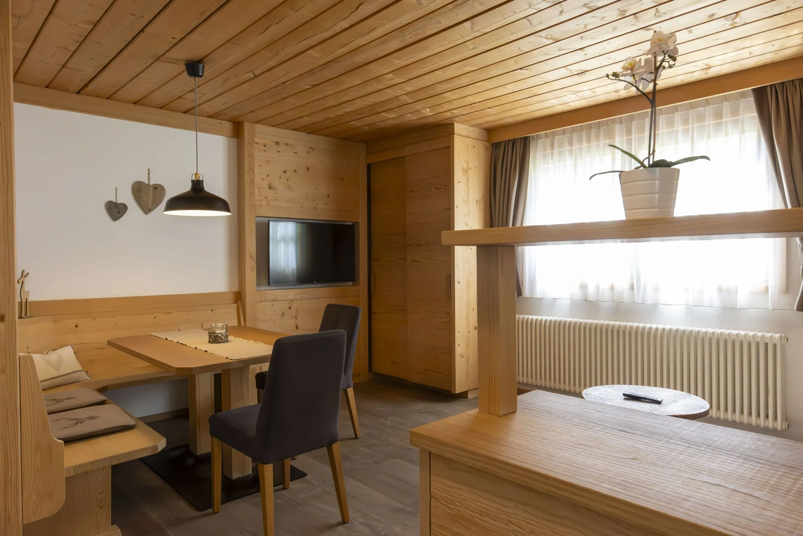 Cozy wooden dining area with bench seating, table, TV, and a windowed corner planter in a rustic interior.