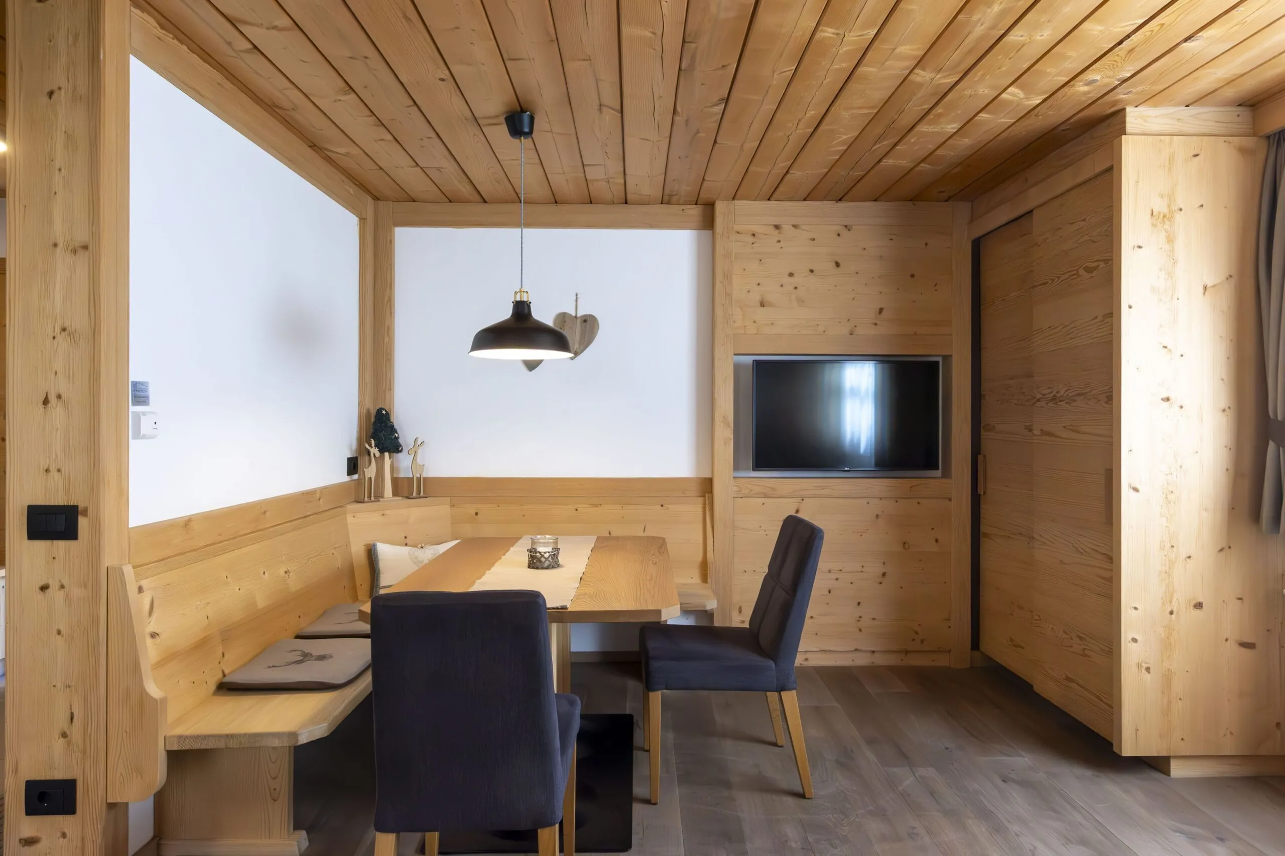 Cozy wooden dining nook with bench seating, a rectangular table, navy chairs, and a mounted flat-screen TV.