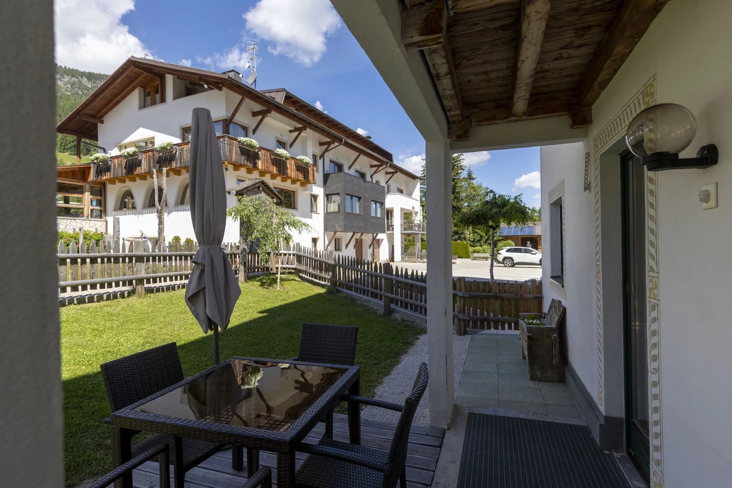 Outdoor hotel courtyard with wooden tables, chairs, umbrella, fenced lawn and white alpine building across.