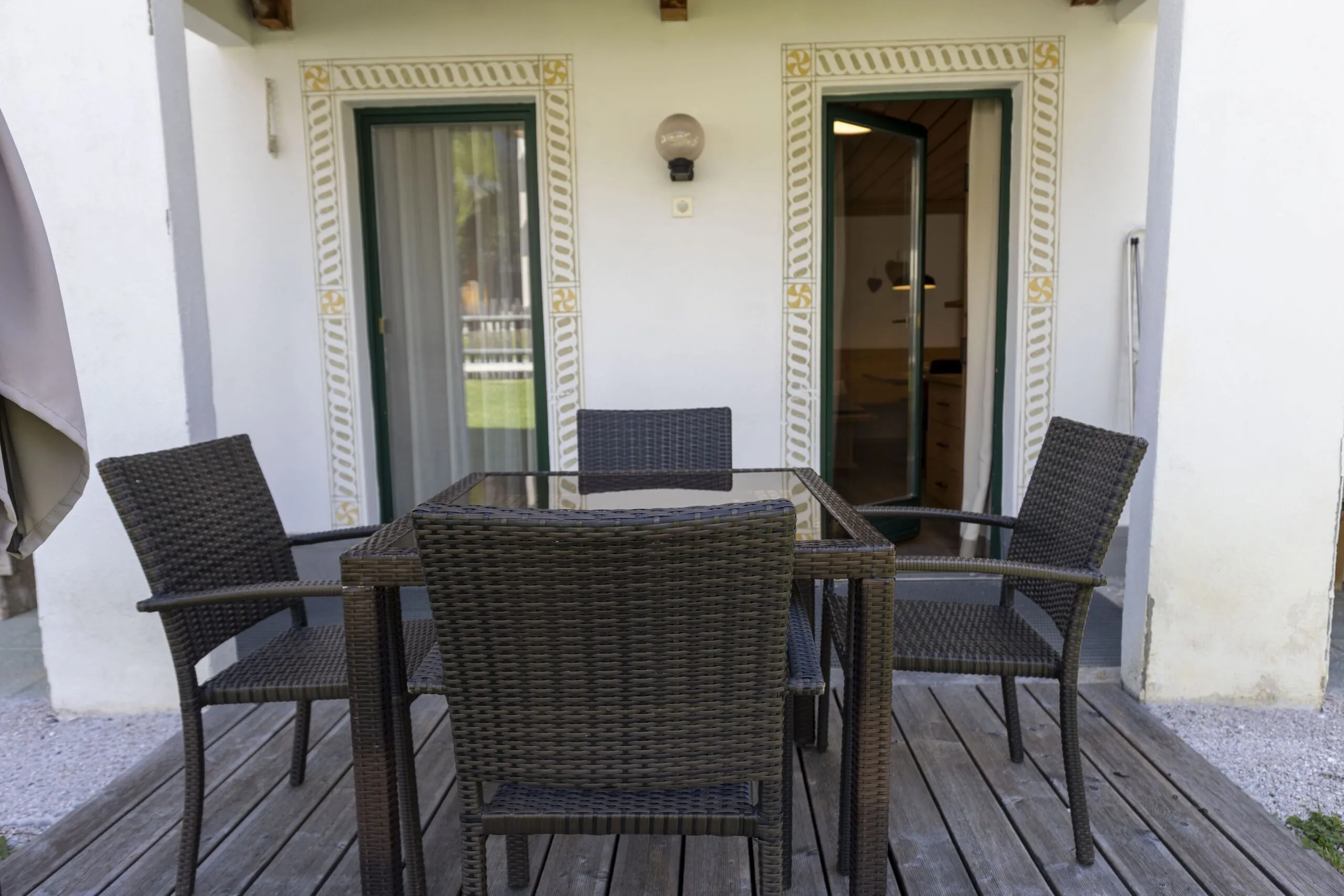 Outdoor patio with dark wicker dining set on wooden deck in front of a white wall with decorative trim.