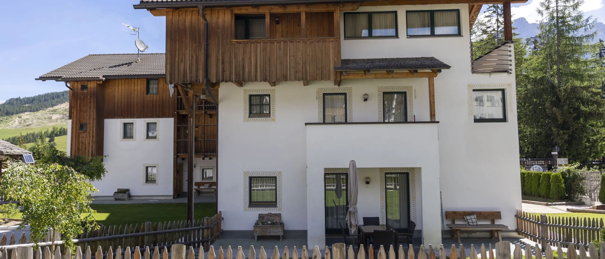 Charming white and wood alpine building with balcony, patio, and surrounding fence in a green mountainous setting.