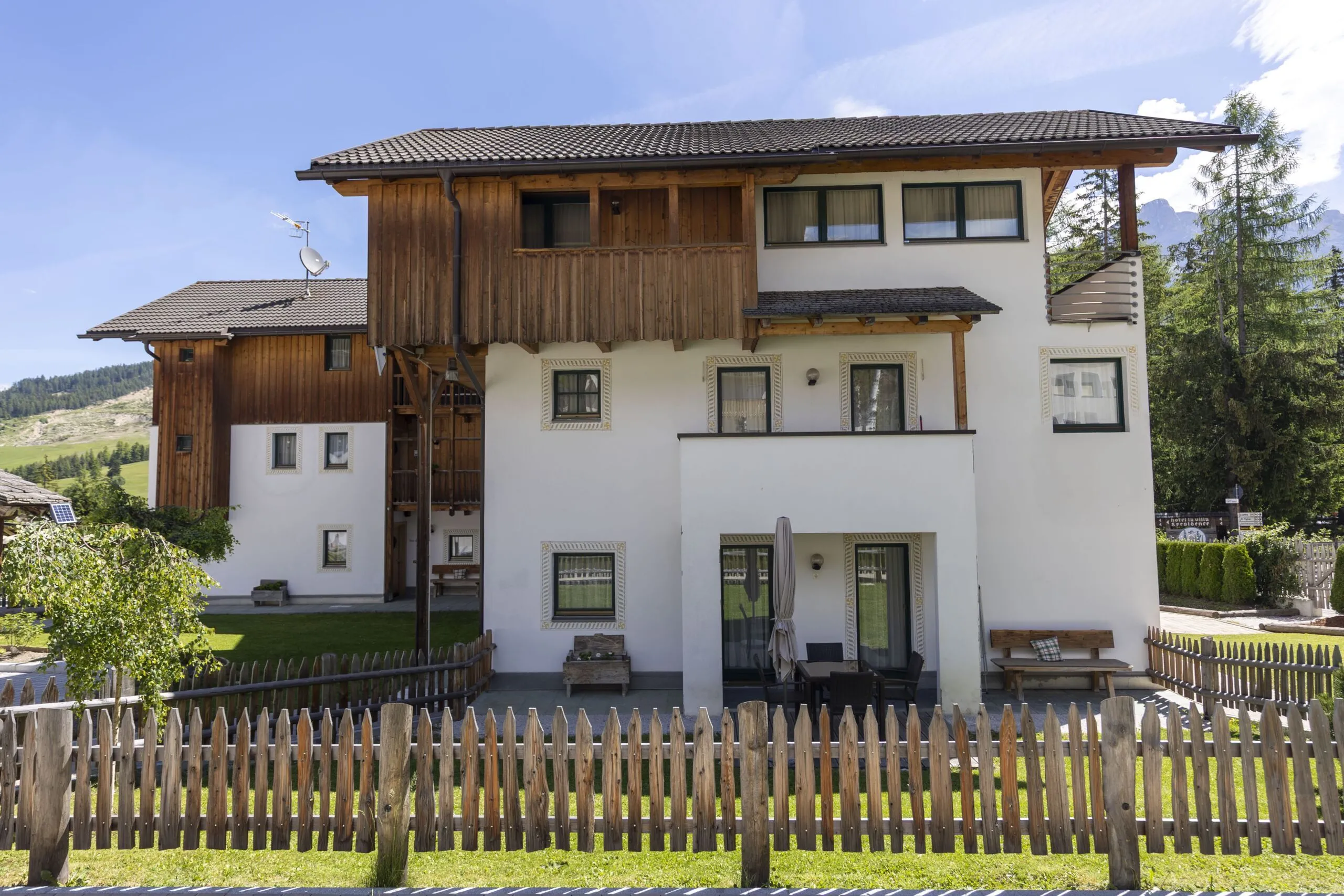 Charming white and wood alpine building with balcony, patio, and surrounding fence in a green mountainous setting.