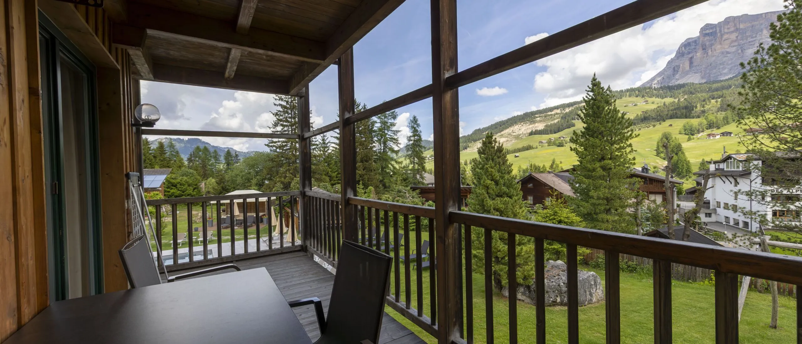 Balcony with wooden railing and table facing a scenic alpine village and green hills under a blue sky.