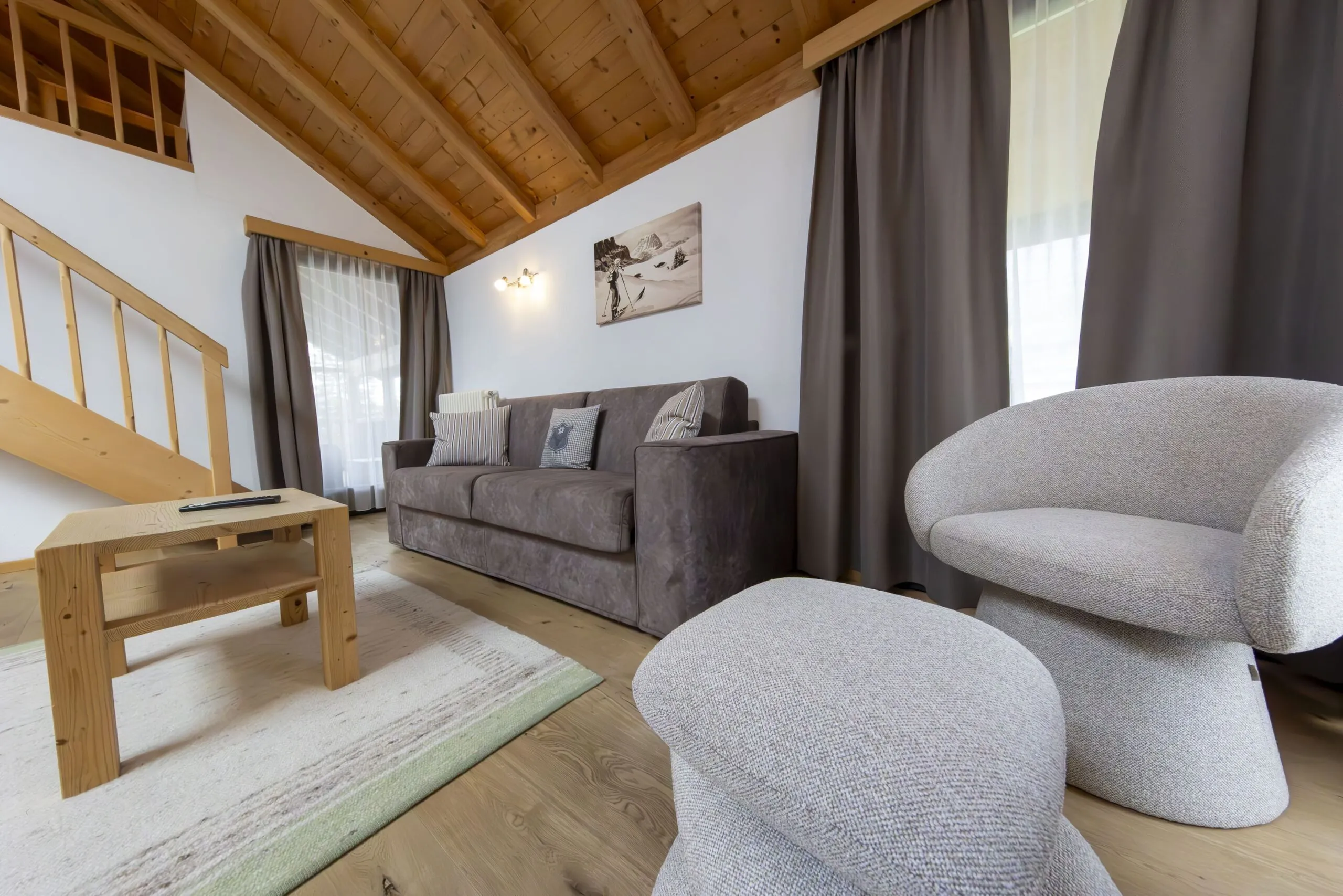 Cozy living room with wooden beams, gray sofa, curved gray armchairs, coffee table, and large windows with curtains under a sloped ceiling.