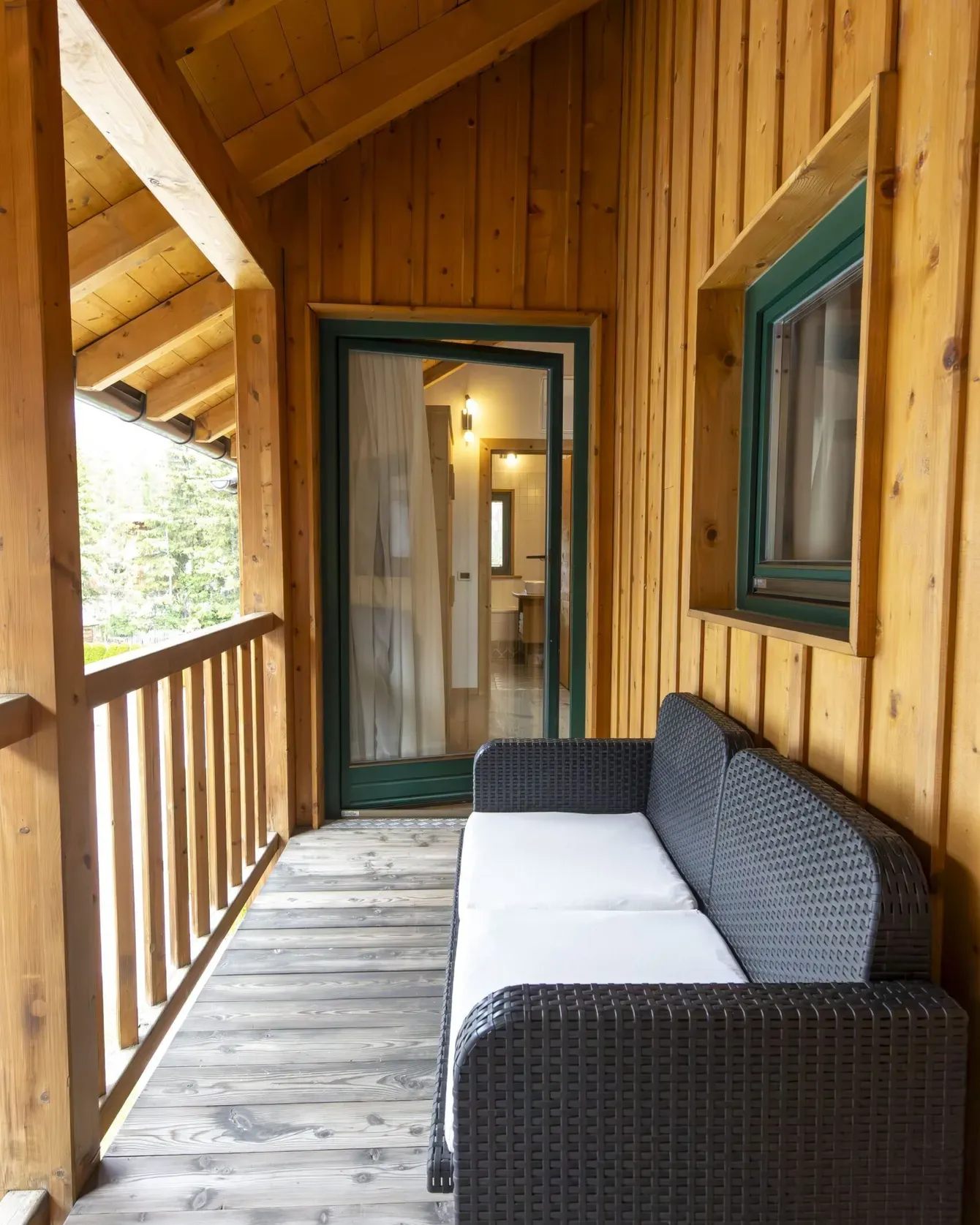 Wooden cabin balcony with wicker sofa and teal-framed door to an interior room, bright outdoors visible.