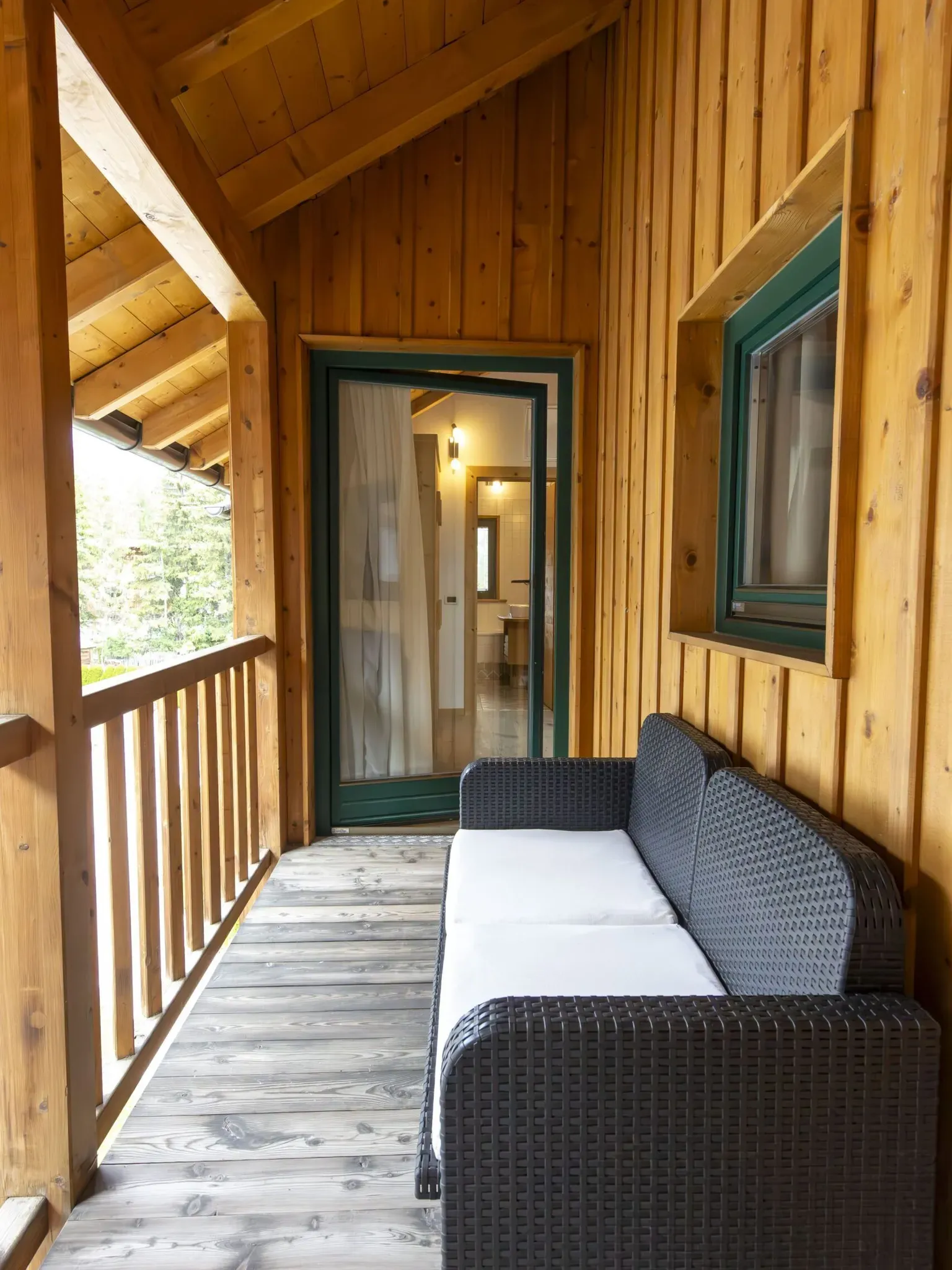 Wooden cabin balcony with wicker sofa and teal-framed door to an interior room, bright outdoors visible.