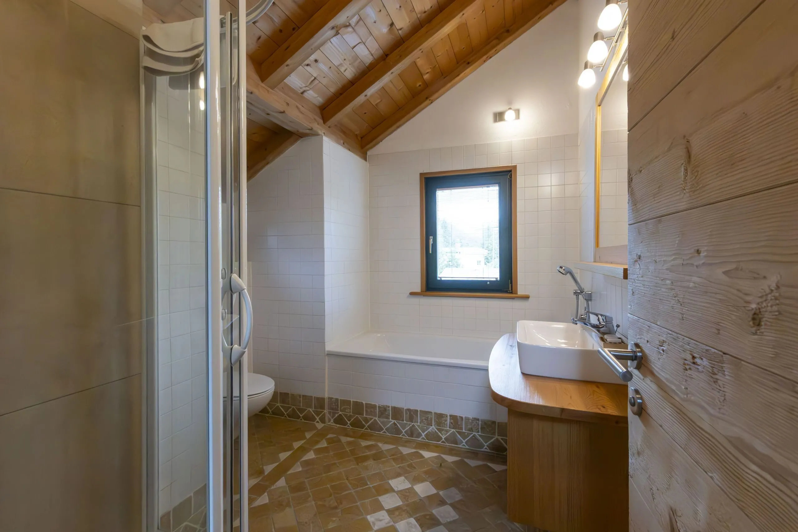 Bathroom with wooden ceiling, white tiled walls, window, white sink, and glass shower enclosure.