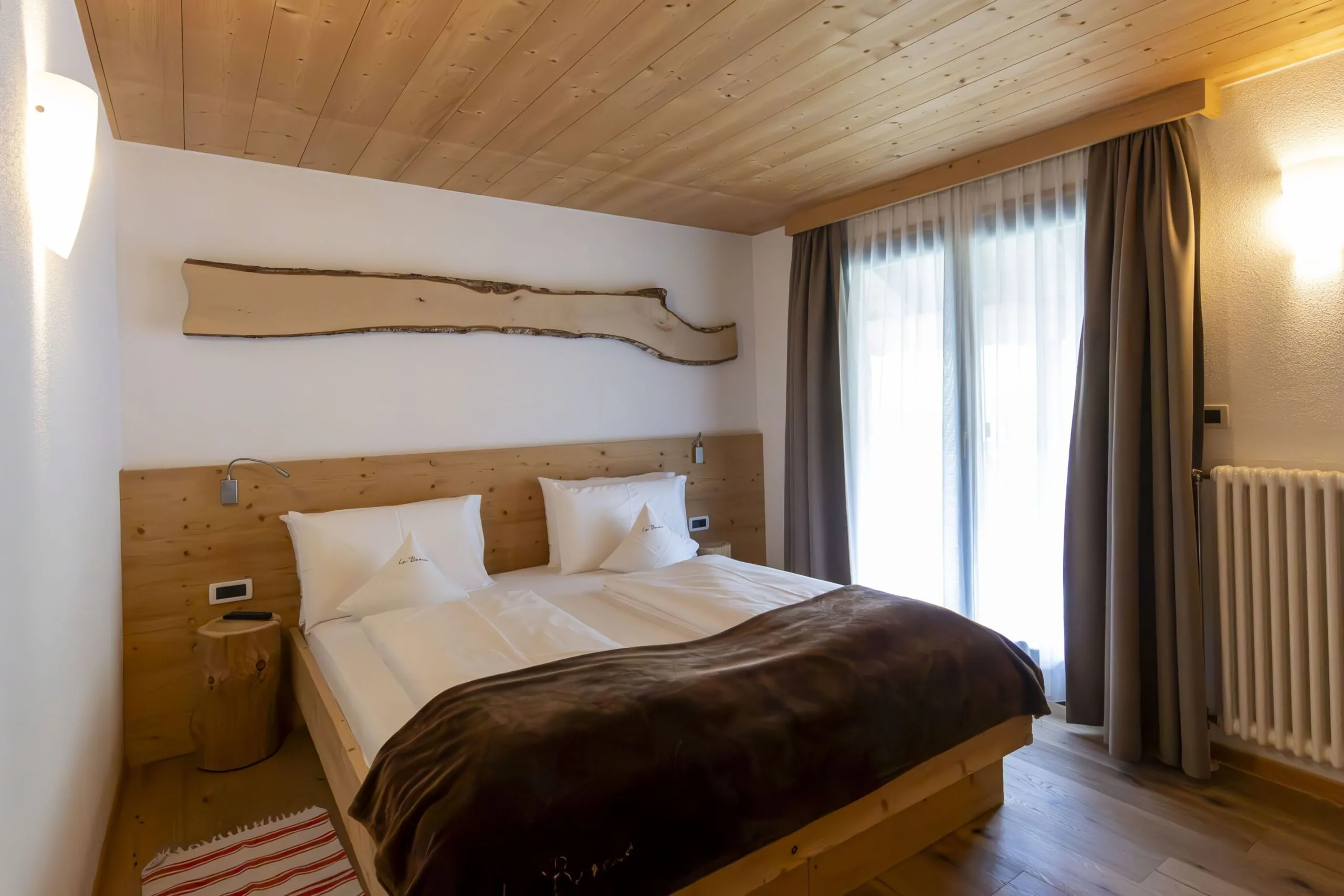 Bright alpine hotel room with a wooden bed, white linens, brown throw, natural wood headboard, and large window with sheer and dark curtains.