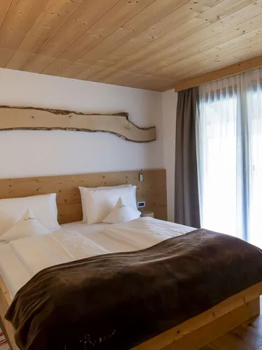 Bright alpine hotel room with a wooden bed, white linens, brown throw, natural wood headboard, and large window with sheer and dark curtains.