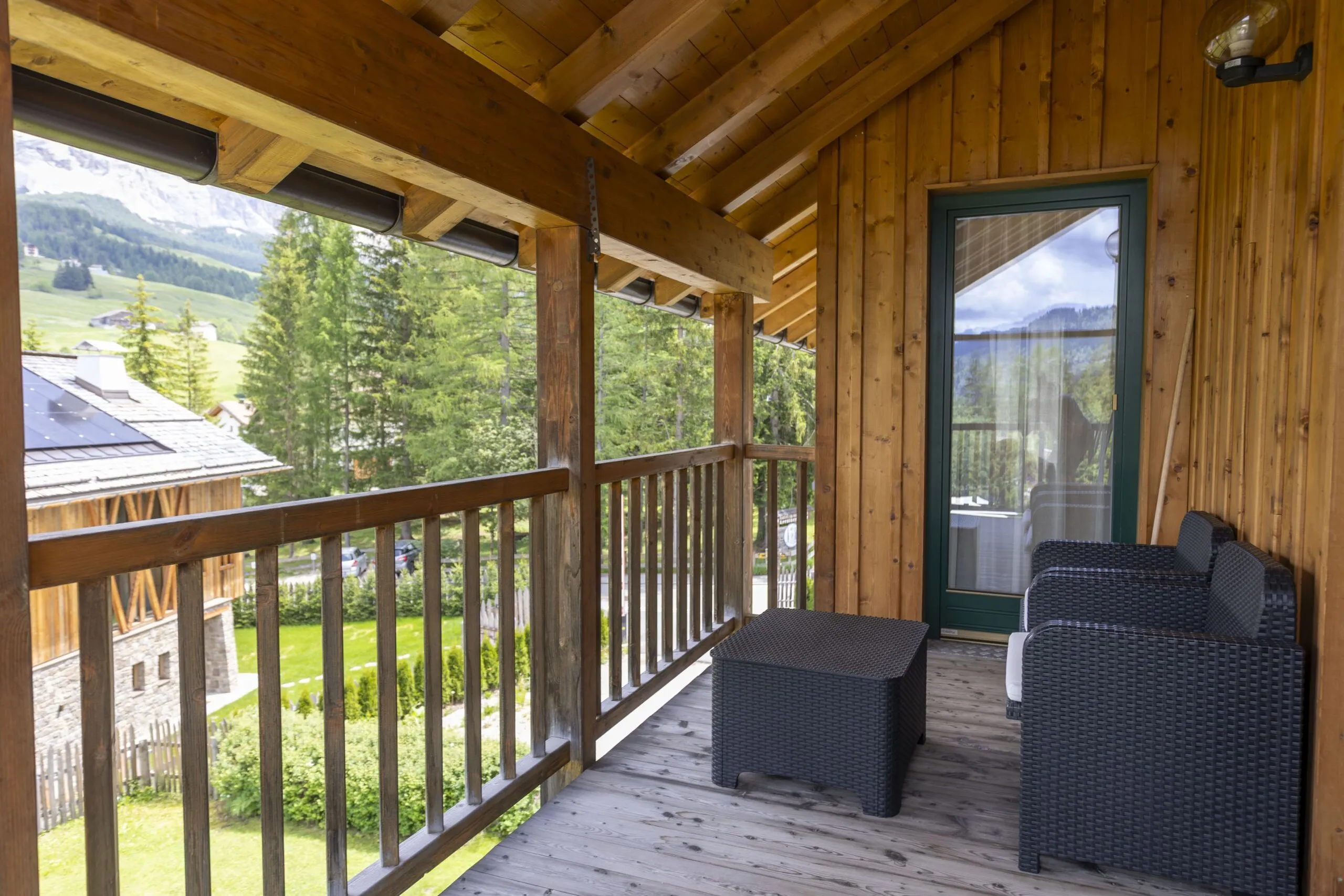 Cozy wooden balcony with black wicker chairs and table, overlooking green valley and trees.