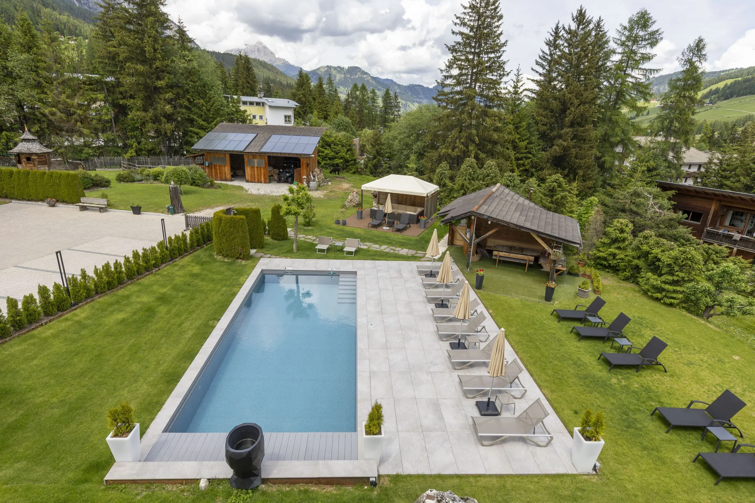 Aerial view of a villa garden with a rectangular pool, sun loungers, shaded seating areas, and surrounding trees.