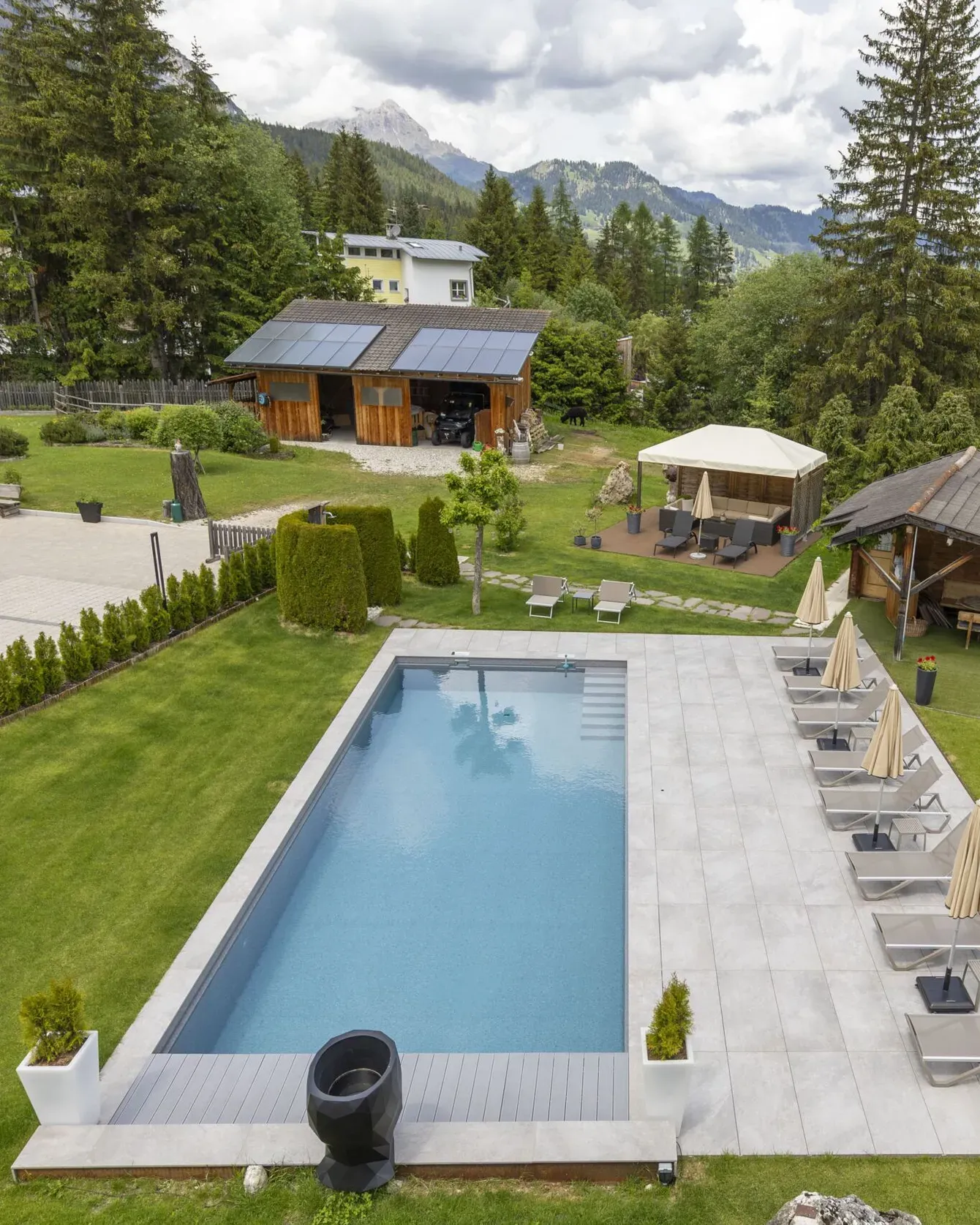 Aerial view of a villa garden with a rectangular pool, sun loungers, shaded seating areas, and surrounding trees.