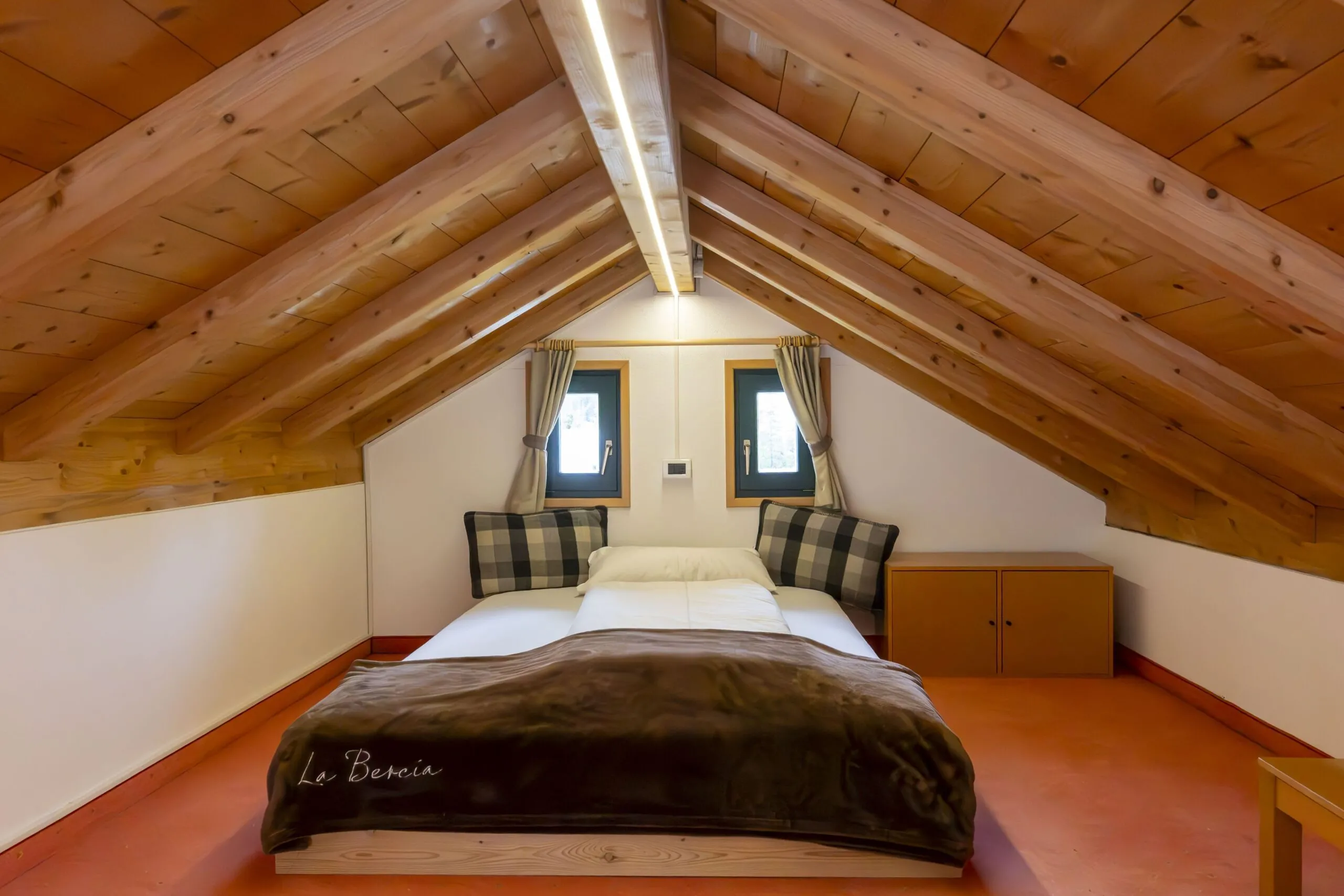 Cozy attic bedroom with a double bed, plaid pillows, and wooden sloped ceiling beams.