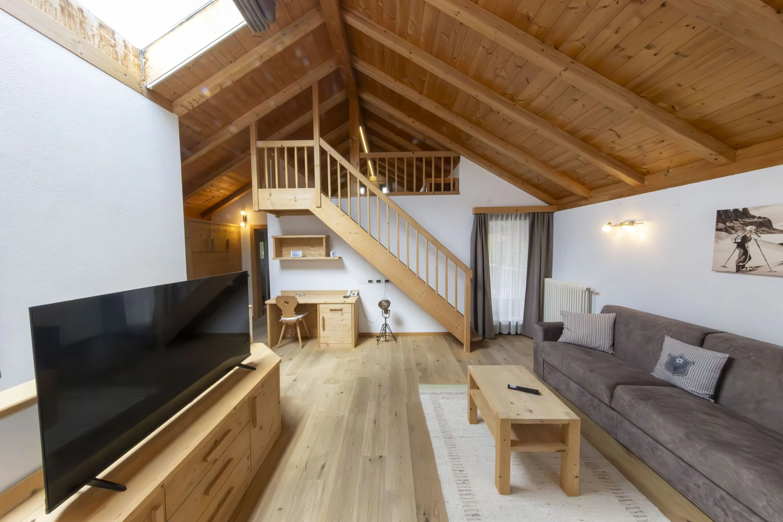 Bright open living area with a wooden loft, large TV, gray sofa, and rustic desk under sloped ceiling.