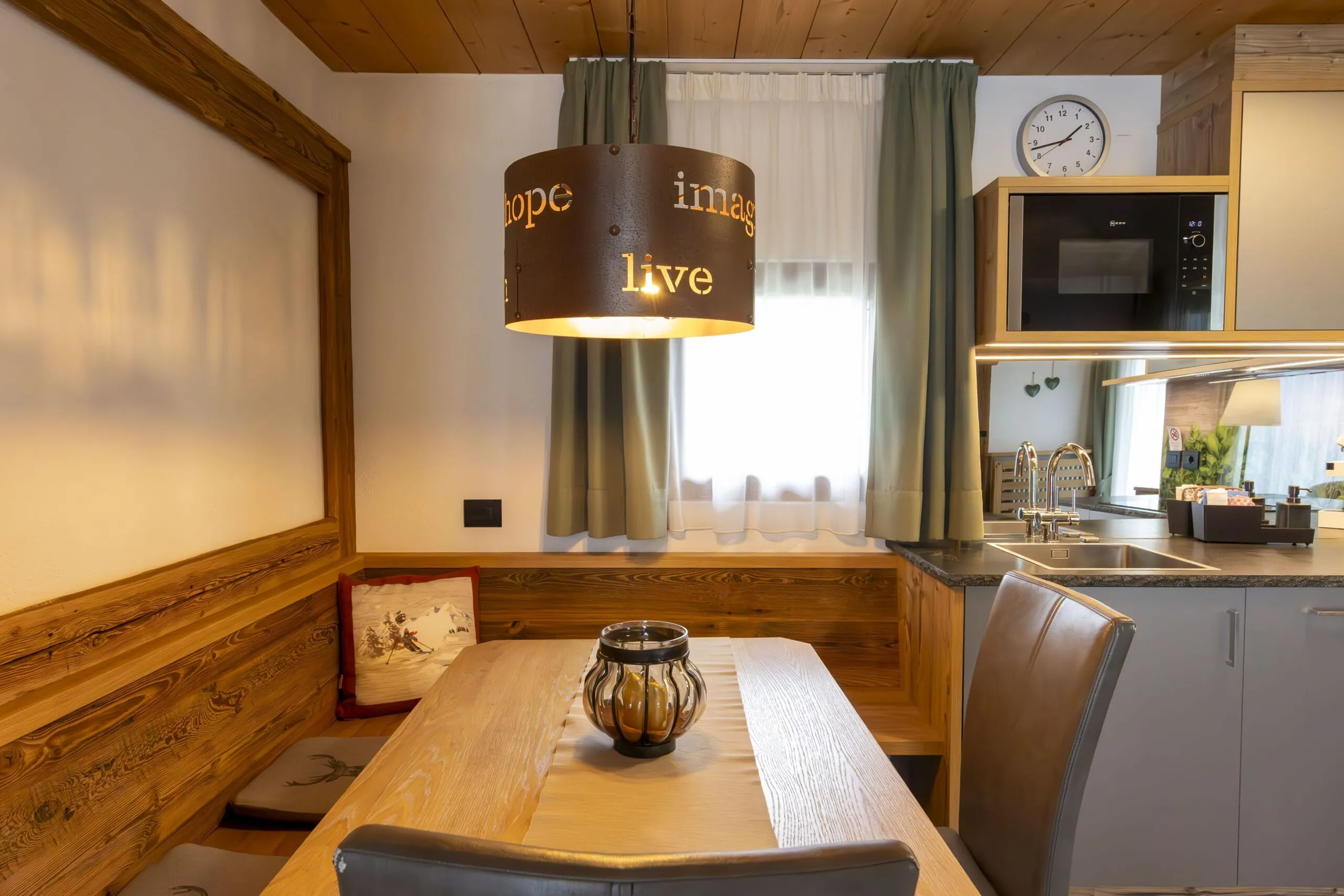 Cozy kitchen-dining nook with wooden benches, a round pendant light, window with green curtains, and a counters kitchen area.