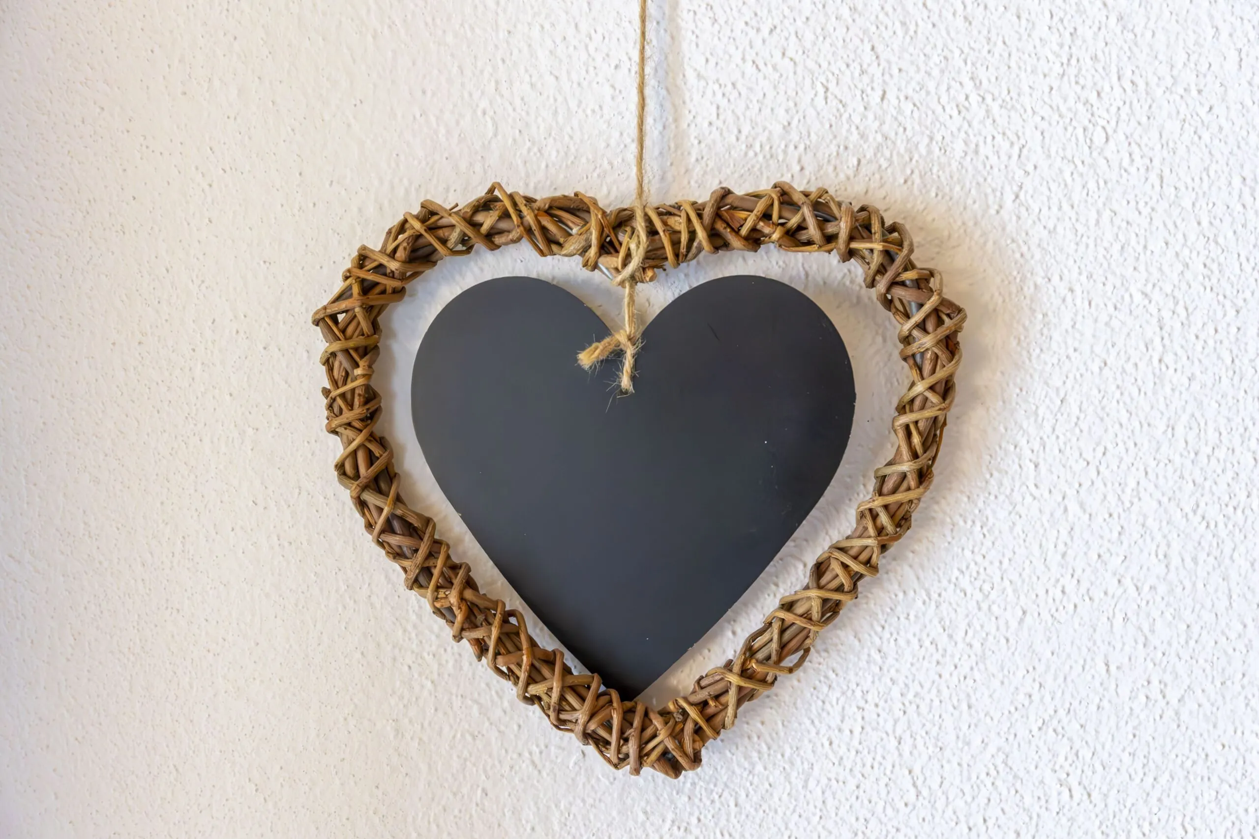 Heart-shaped wicker frame with a dark chalkboard inner heart hanging by jute on a textured light wall.