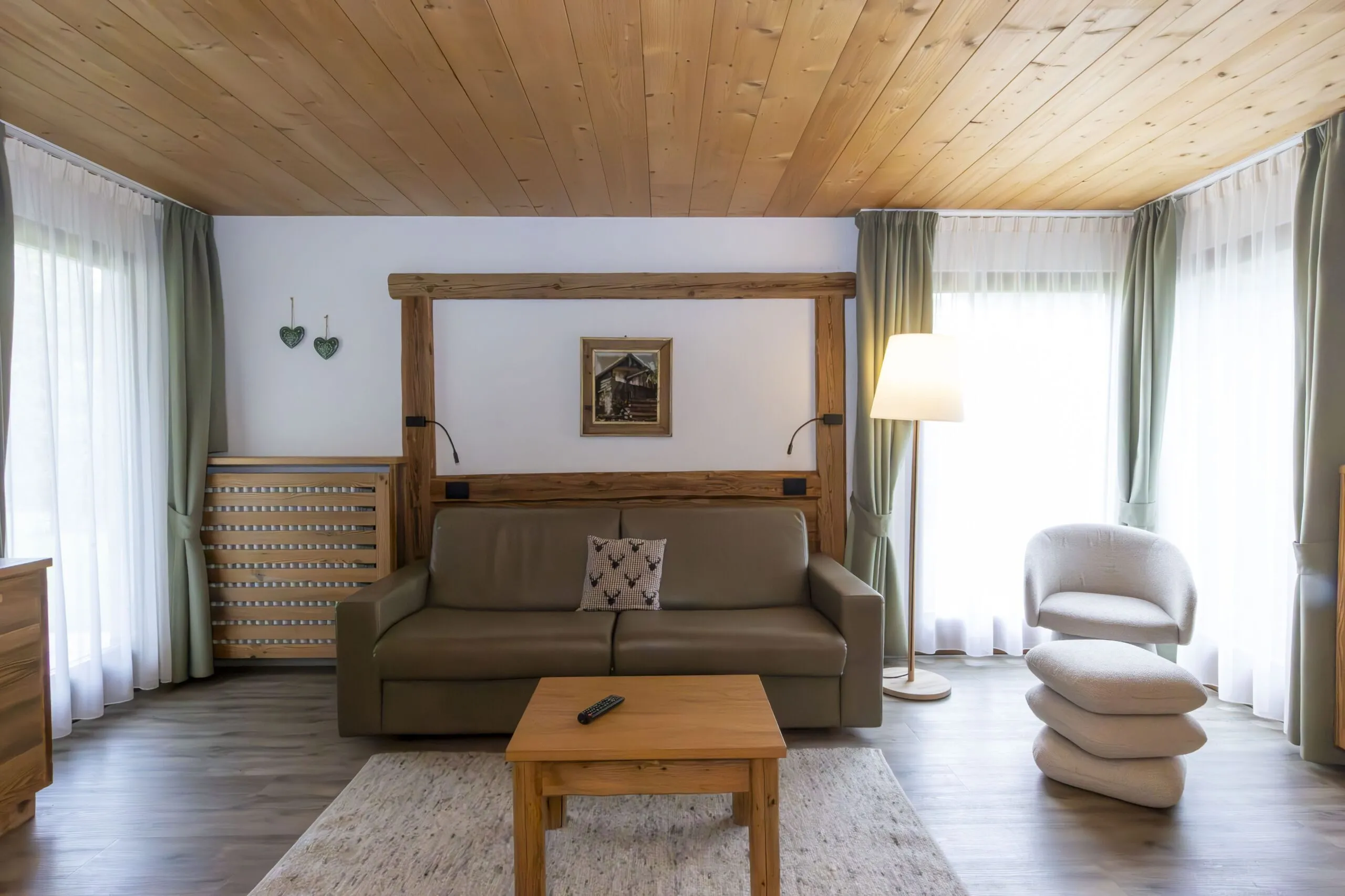Cozy living room with wooden ceiling, beige sofa, light wood furniture, and tall windows with green curtains.