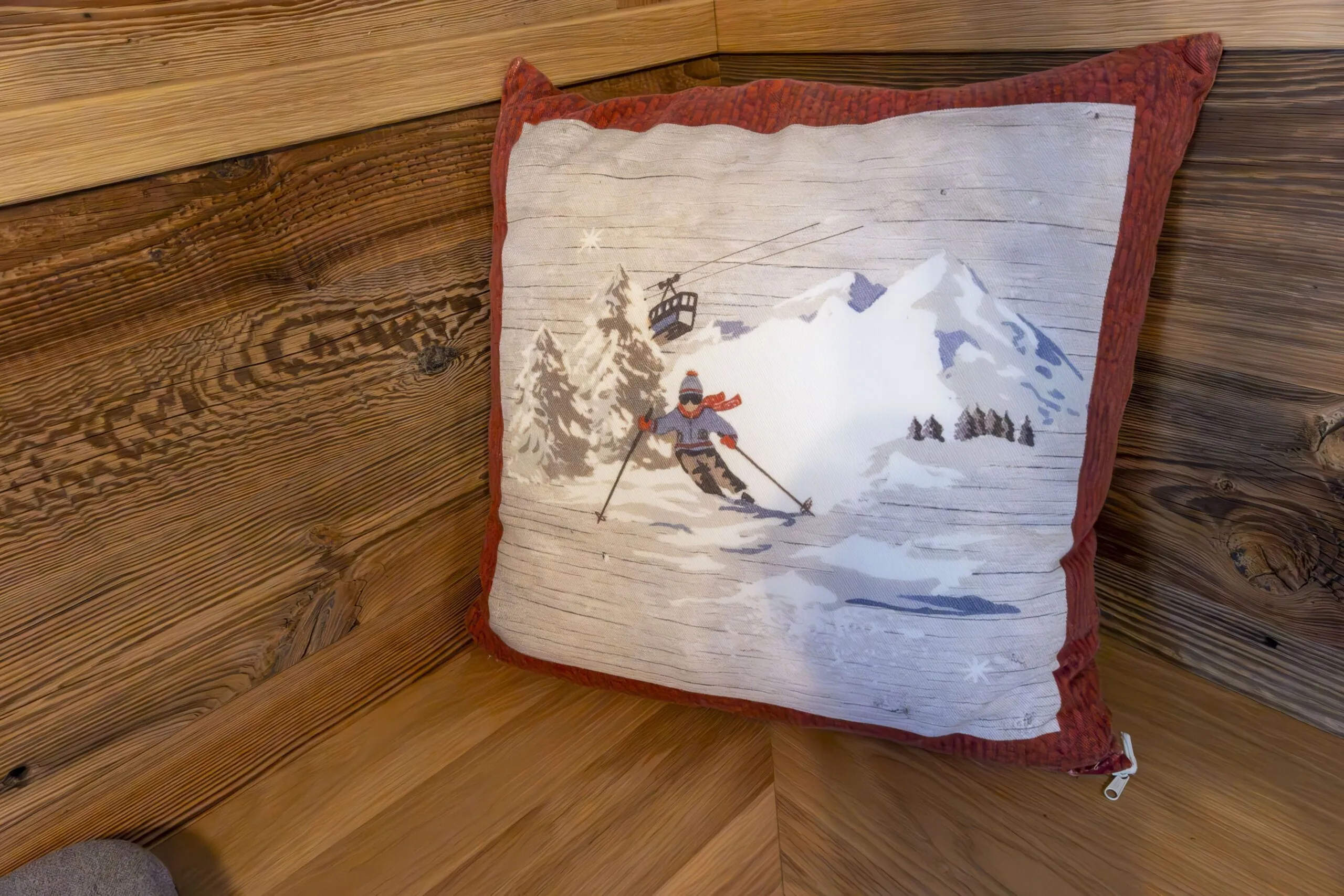Snow skier in blue jacket on slope with mountains and lift in snowy alpine scene on a red-framed cushion.