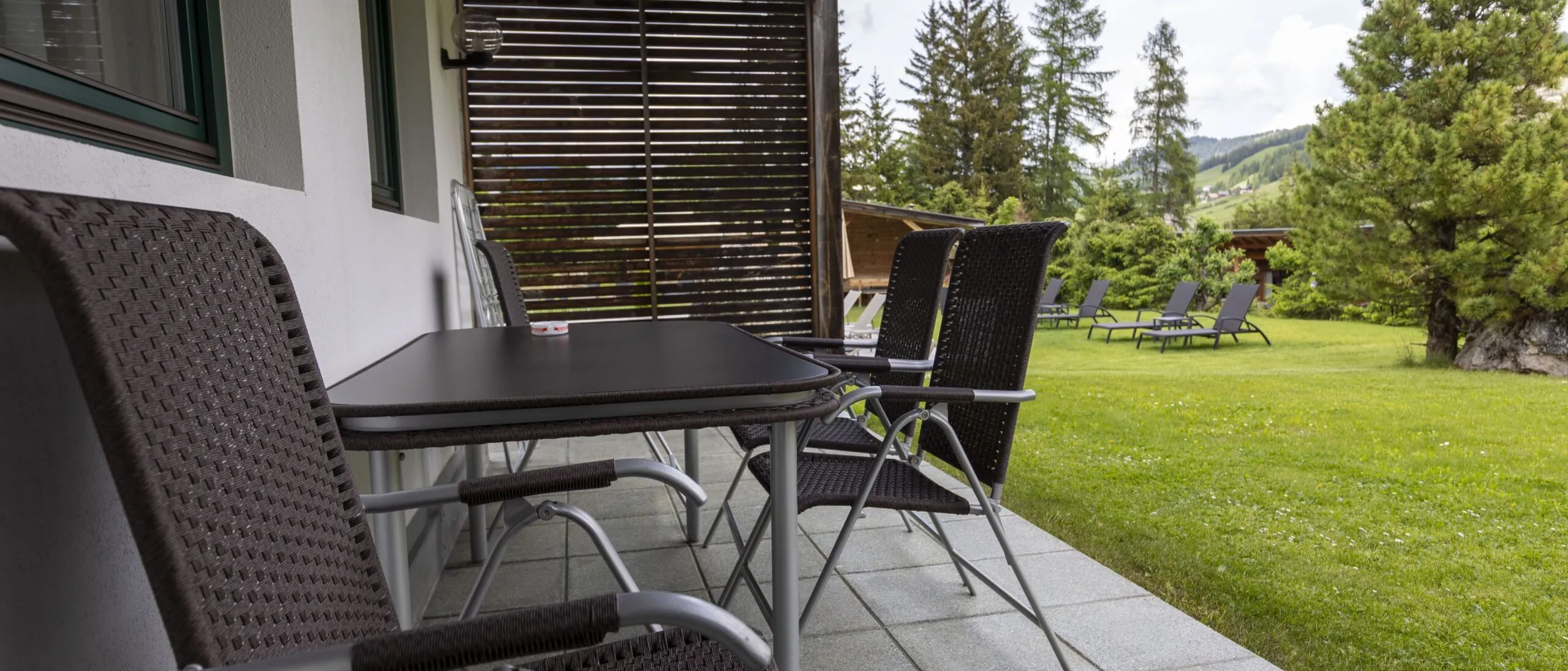 Patio with brown wicker chairs and a black table along a white wall, overlooking a green lawn and trees.