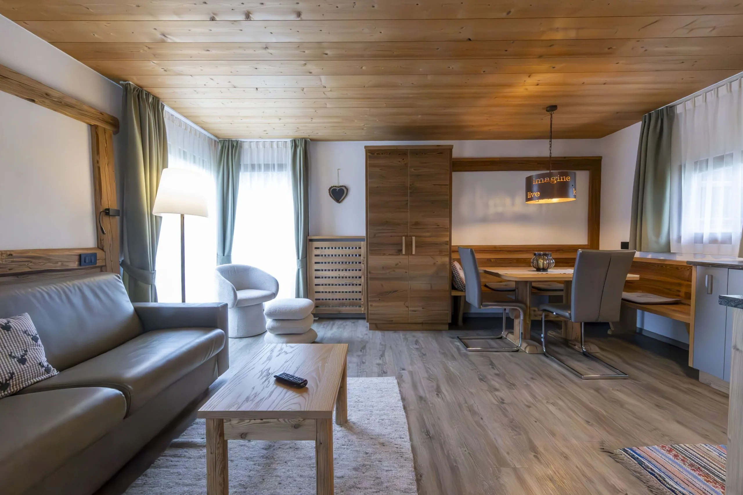 Cozy living area with wooden ceiling and furniture, gray sofa, circular chair, dining nook, and large windows with curtains.