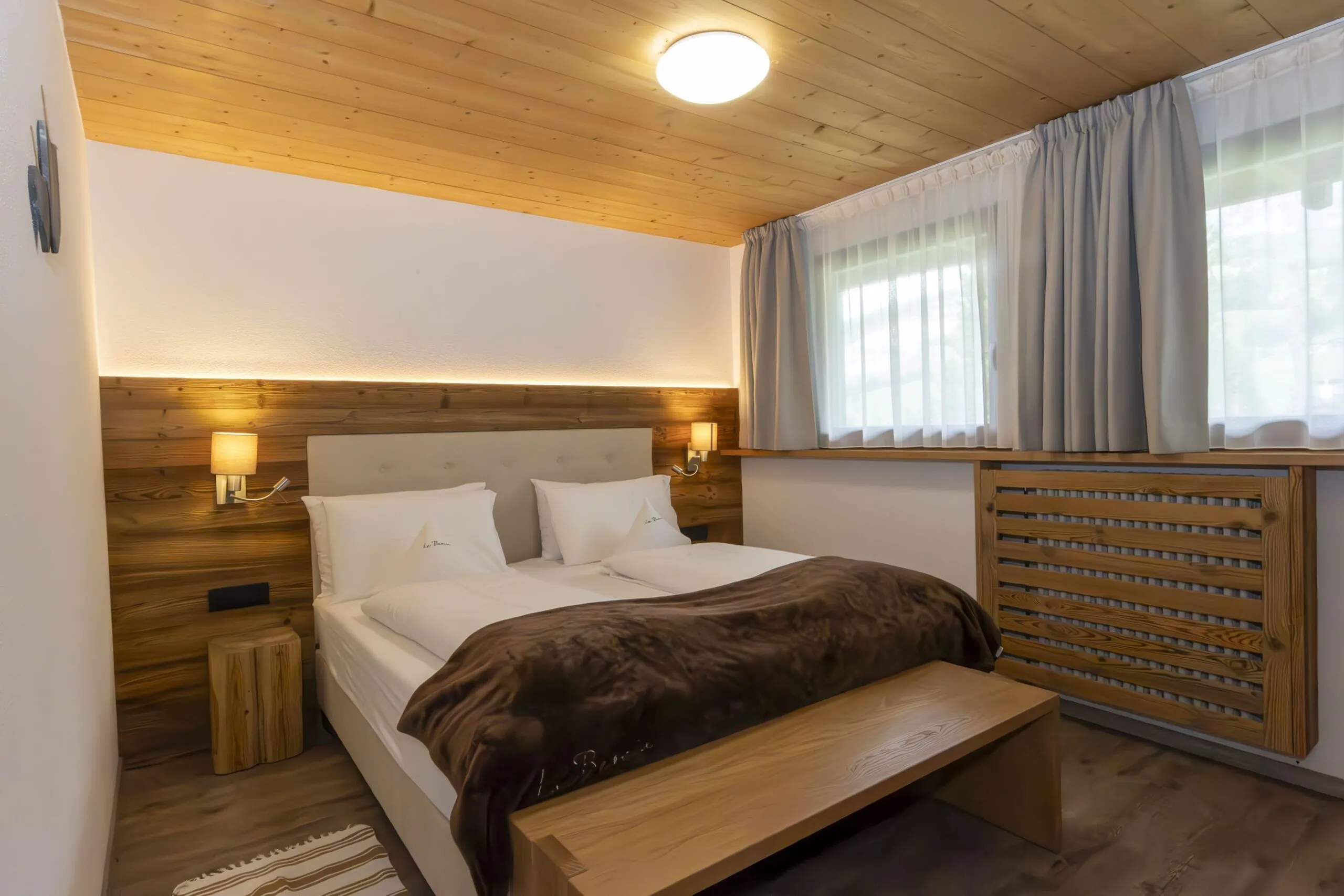Cozy hotel bedroom with a wooden headboard, two white beds, brown blanket, nightstands, and large window with light curtains