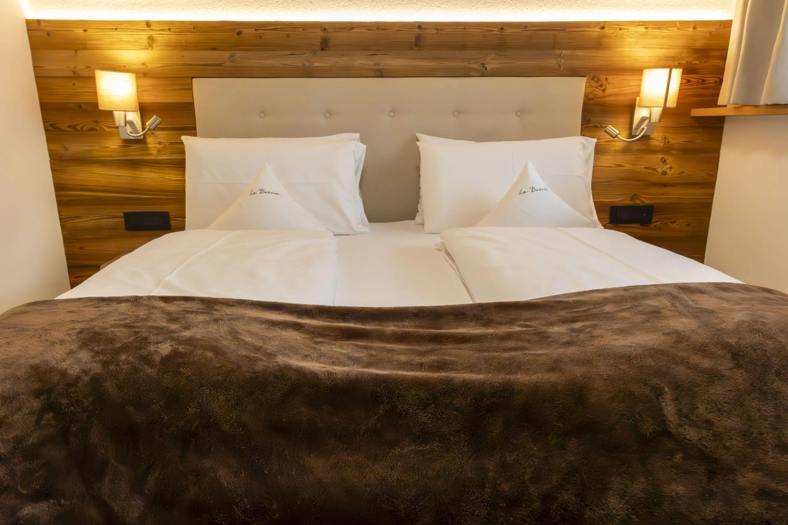 Luxurious twin bed with white pillows and a brown faux fur throw in a wood-paneled hotel room headboard area.
