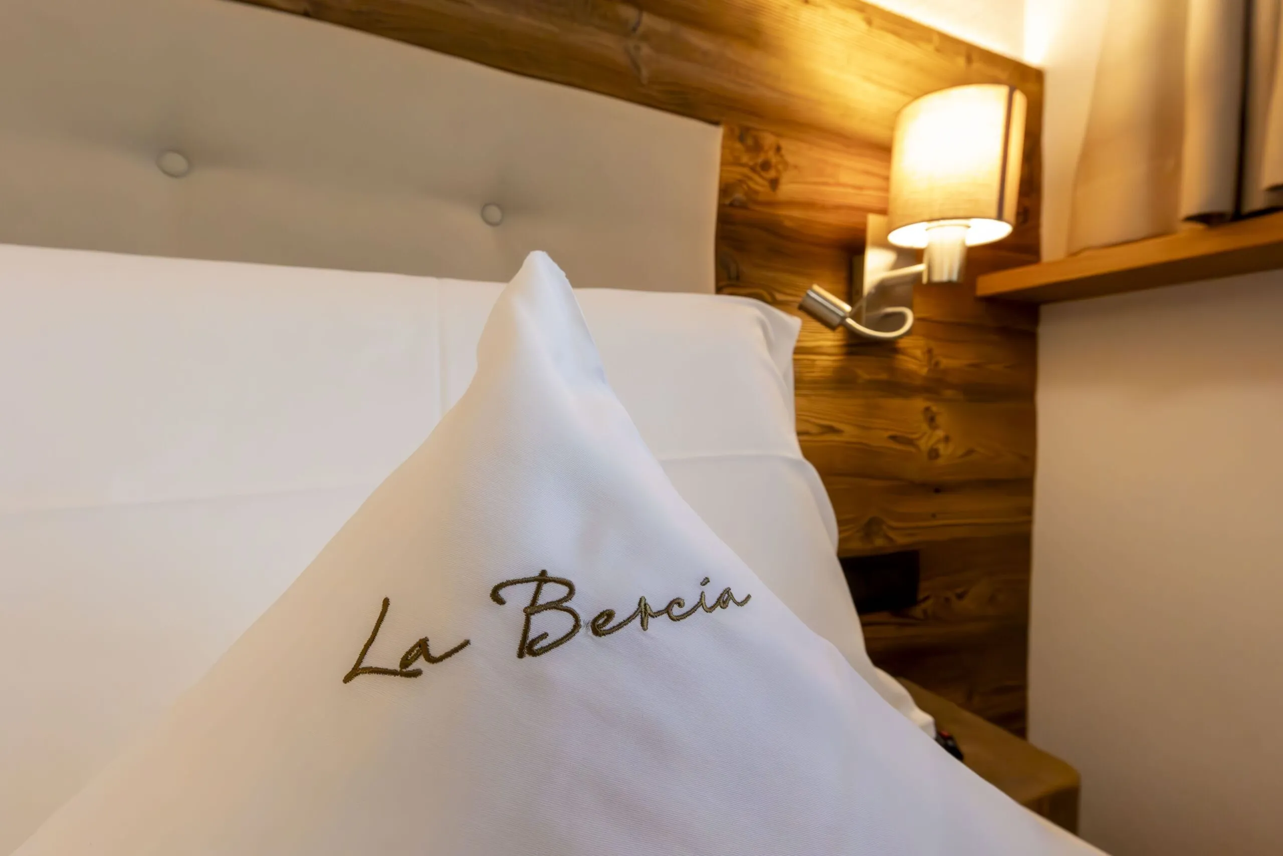 Soft white bed with a folded pillowcase monogrammed “La Bercia” in a cozy wooden hotel room.