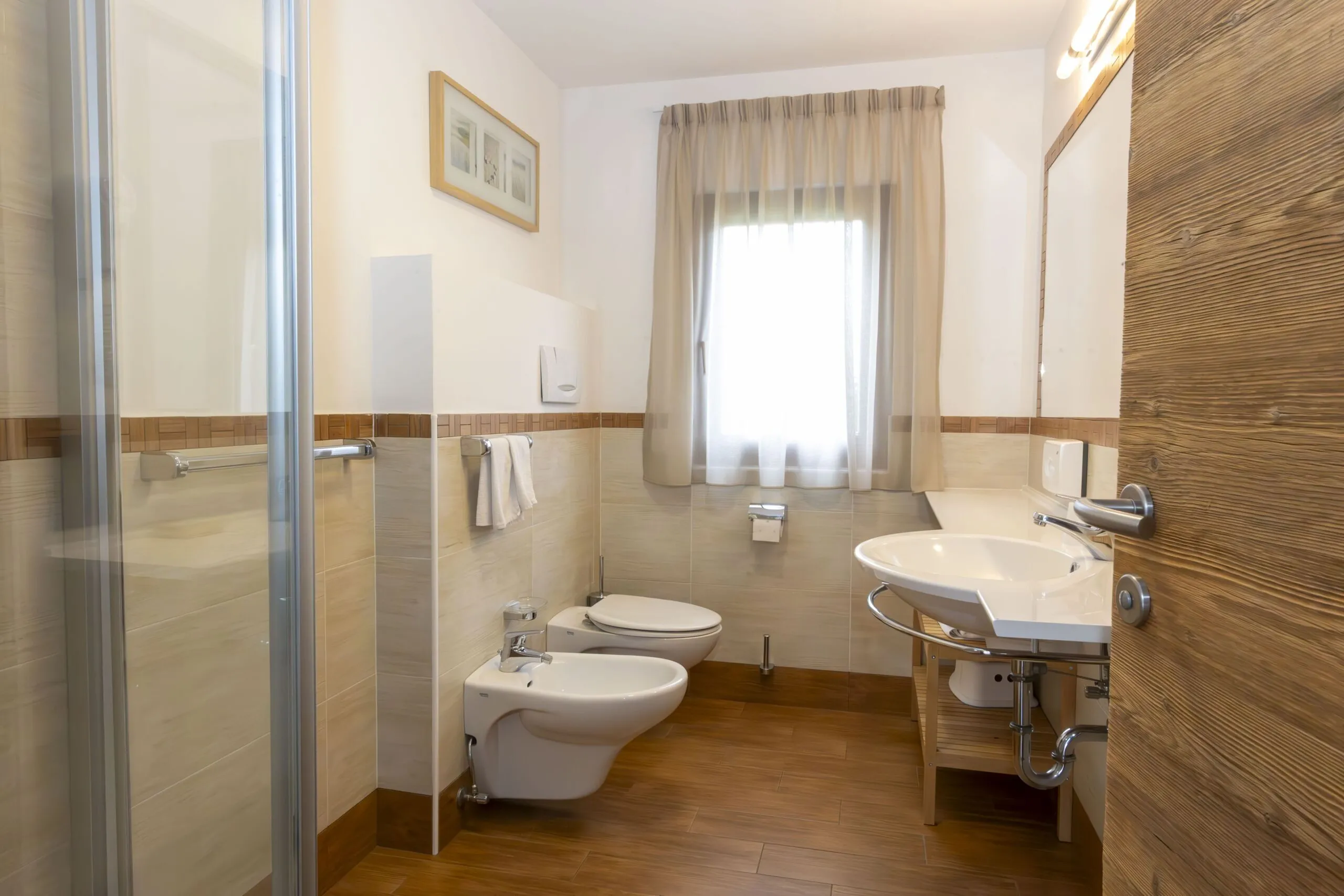 Bright hotel bathroom with window, beige tiled walls, toilet, bidet, sink, wooden floor, and shower enclosure.