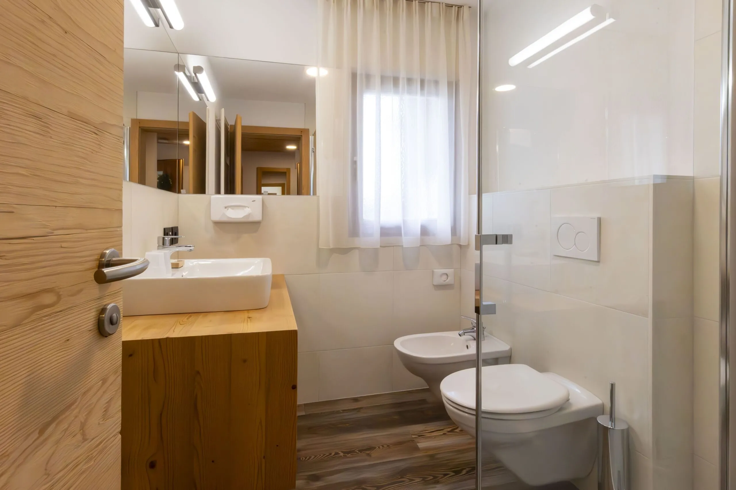 Modern bathroom with wooden vanity, white sink, wall mirror, and glass shower enclosure in bright, minimalist setting.