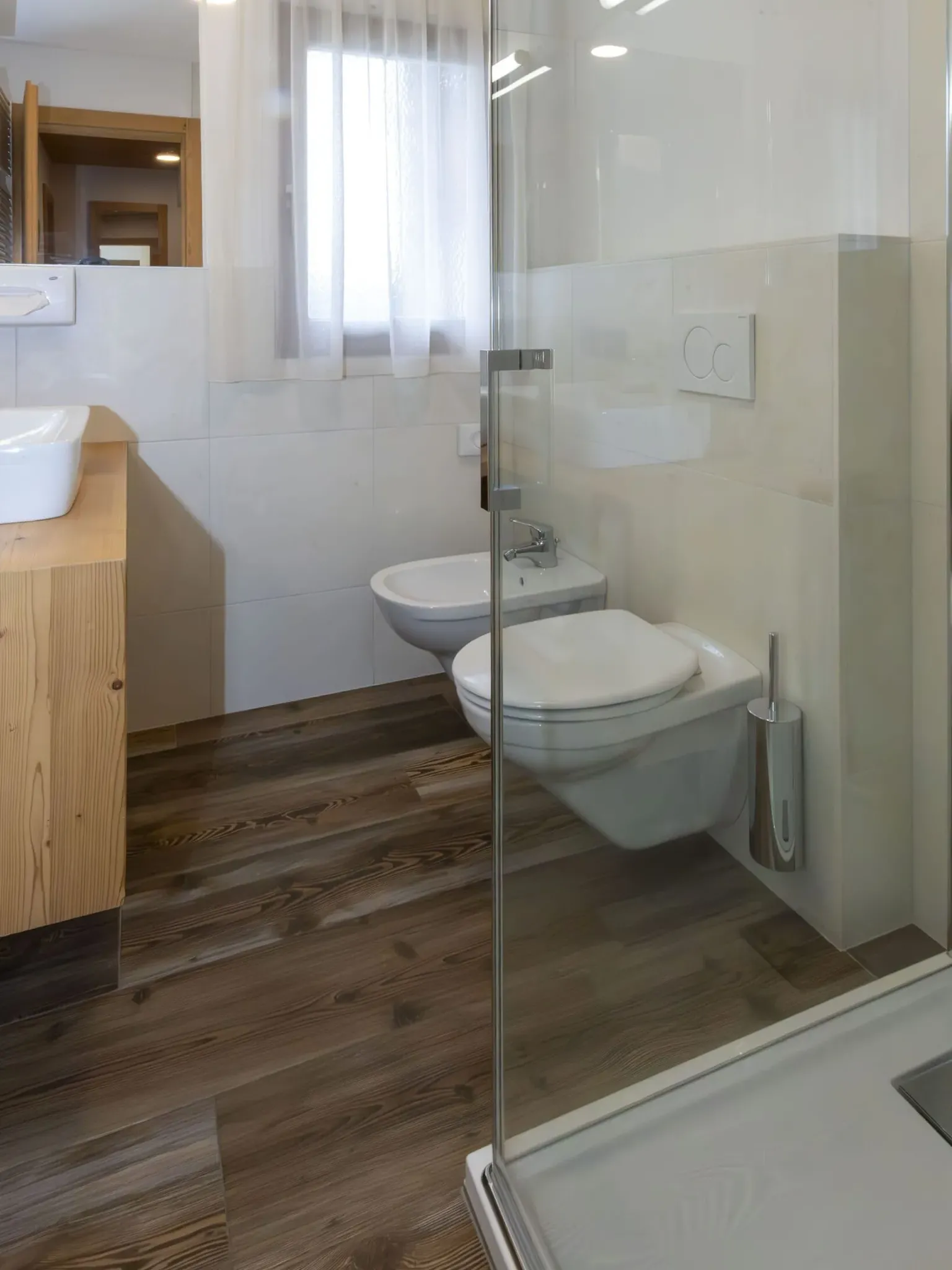 Contemporary bathroom with wall-mounted toilet, bidet, wooden vanity, glass shower, and wood-like flooring