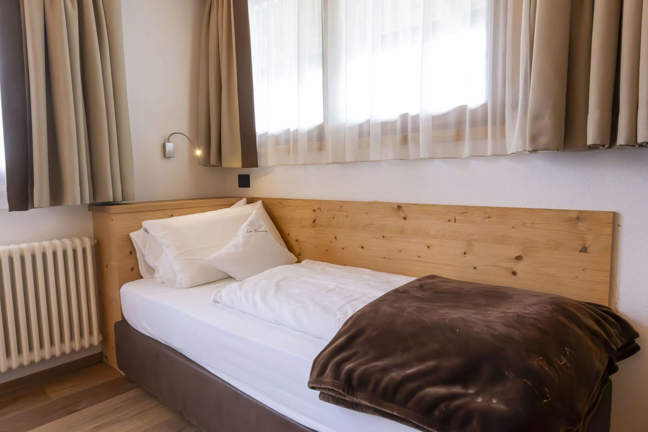 Cozy single bed with white linens and brown blanket, wooden headboard, beige curtains, and wall-mounted reading light.