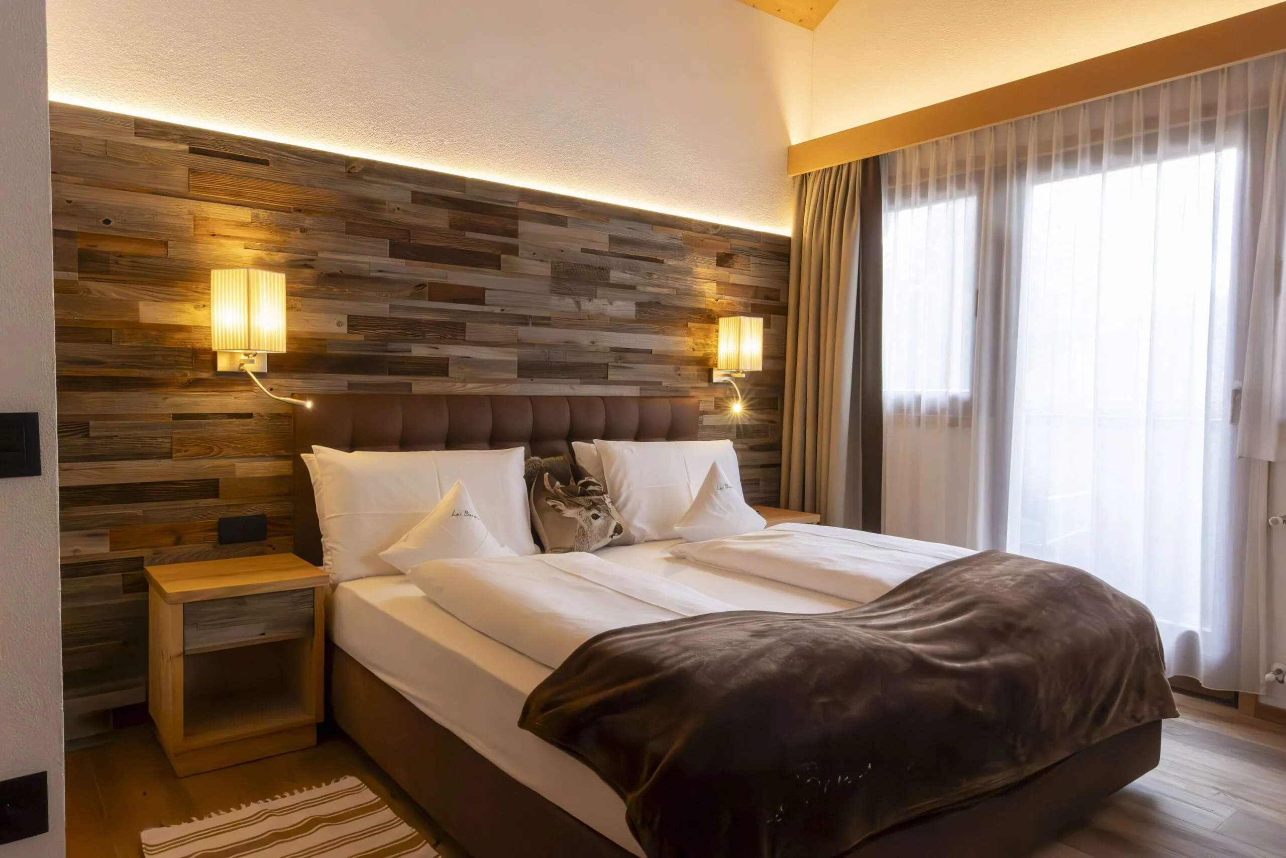 Warm modern hotel room with wooden feature wall, two nightstands, bed with white linens, brown headboard, and sheer-curtained window.
