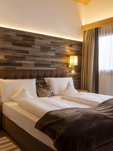 Warm modern hotel room with wooden feature wall, two nightstands, bed with white linens, brown headboard, and sheer-curtained window.